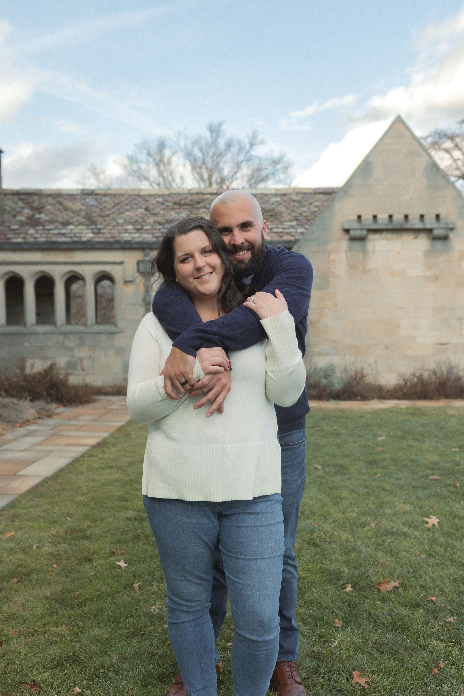 A couple embracing in front of a historic stone building with arched colonnade.