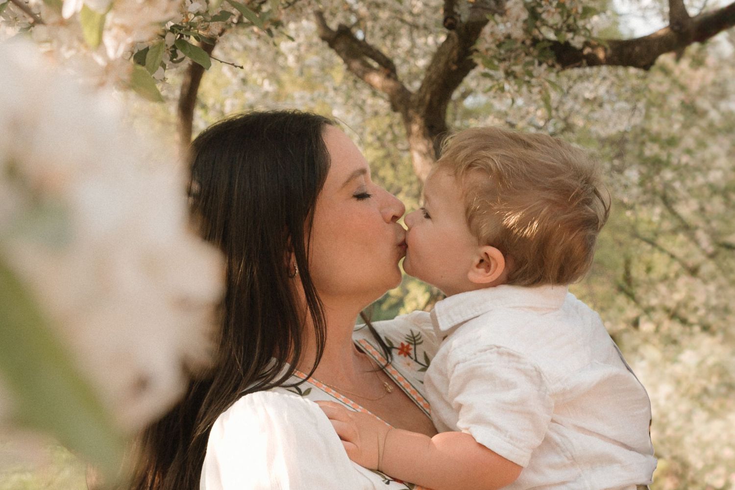 Tender moment of mom and toddler sharing a kiss under blooming trees, spring maternity photo with soft florals and timeless emotion.