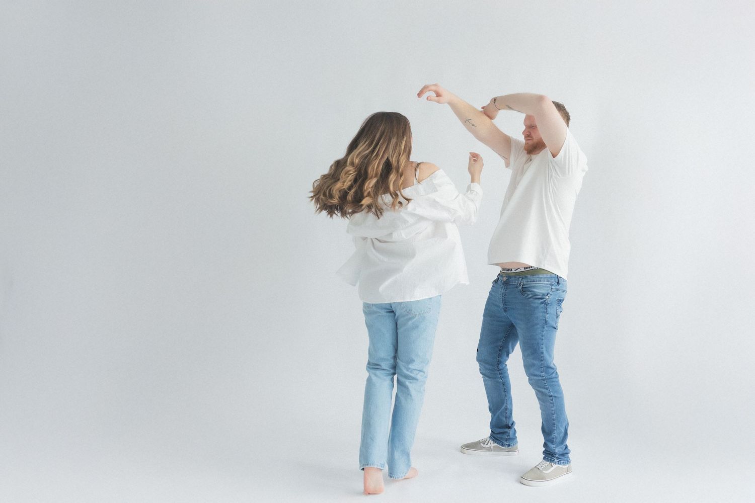 Playful couple dancing together barefoot against white background in matching casual outfits.