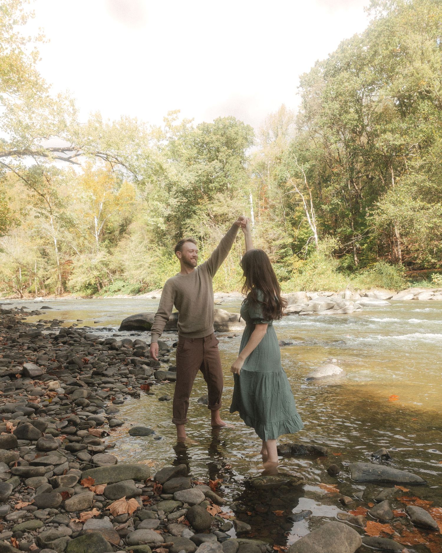 A couple shares a romantic moment by the rocky riverbank on a sunny autumn day.