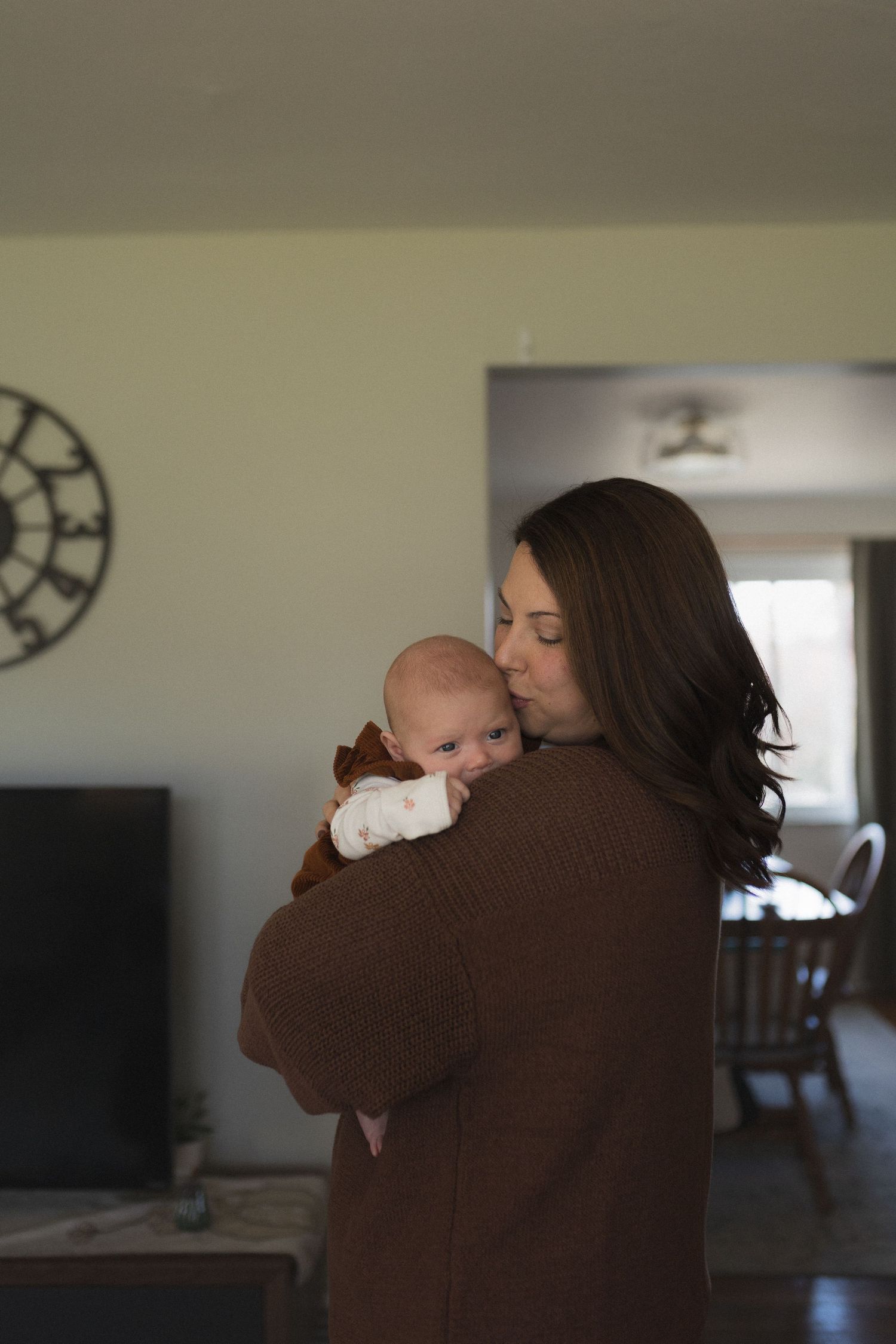 A brown sweater-clad figure holds and comforts a sleeping infant in a living room setting.