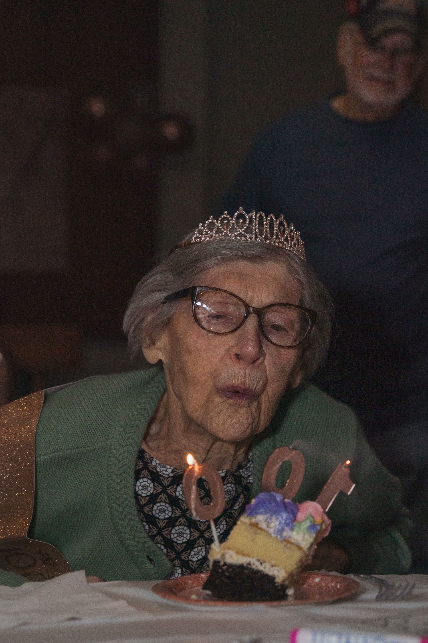 Senior wearing tiara blows out birthday candles on cake.