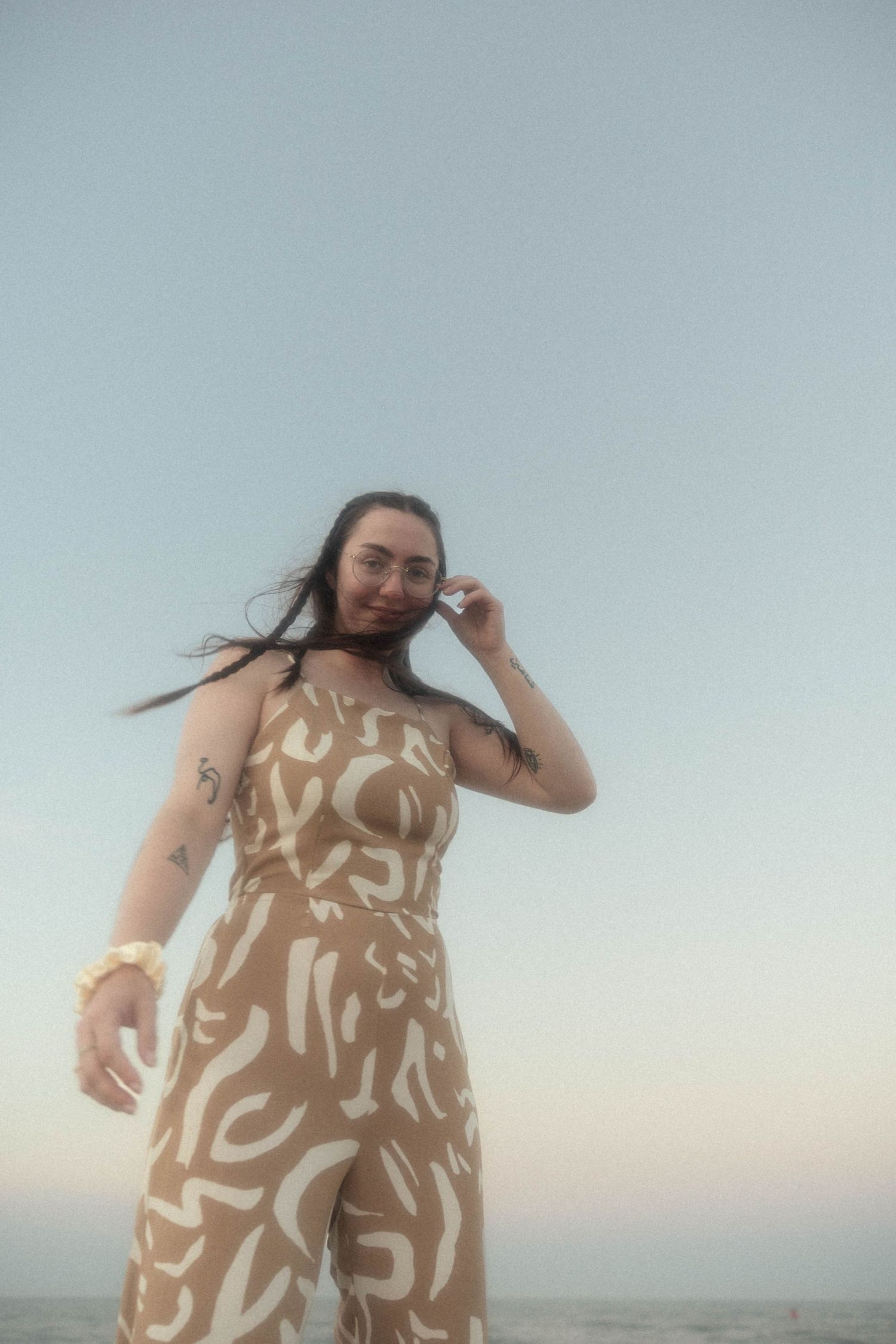 A person in a tan abstract-print dress poses against soft sky at the beach.