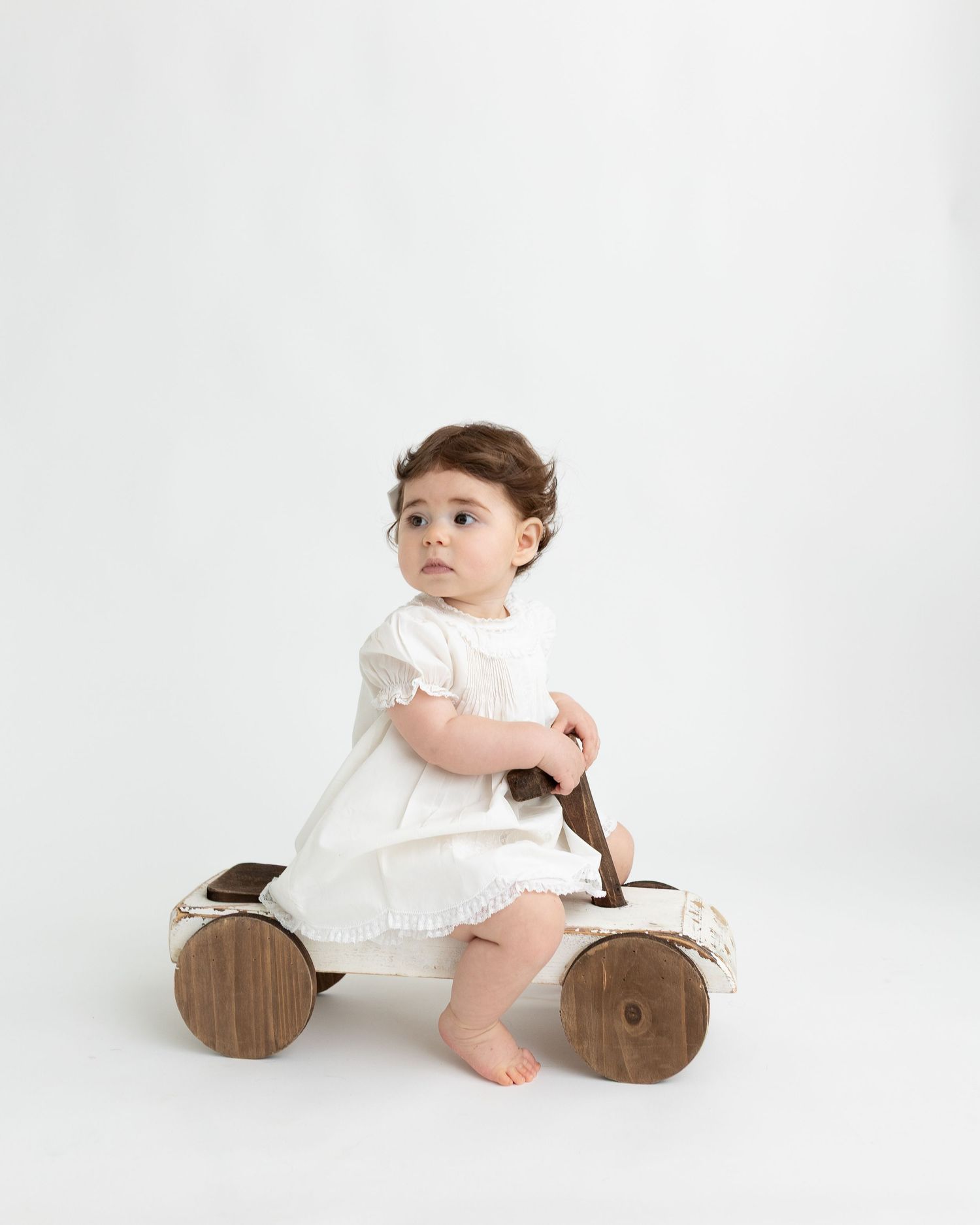 Small child wearing white dress sits on wooden rolling toy against plain white background.