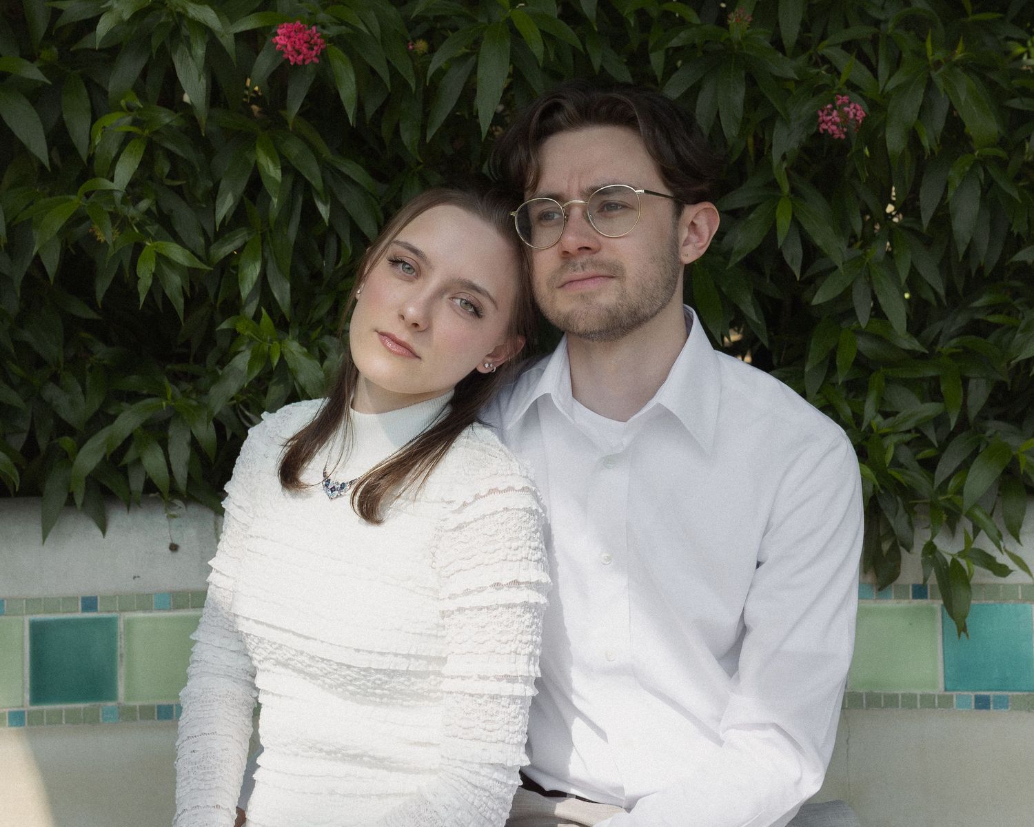A young couple in white attire sitting together outdoors near lush green foliage and a tiled wall.