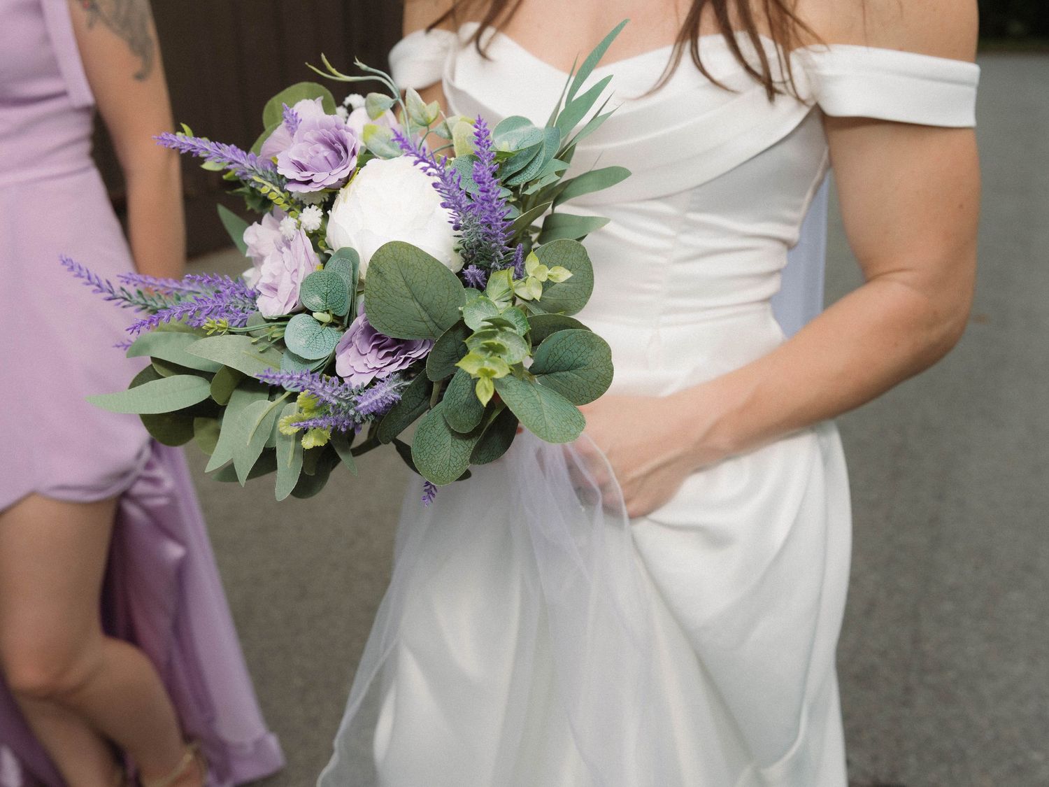 Wedding party members hold purple and white floral bouquets with eucalyptus against their white dresses.