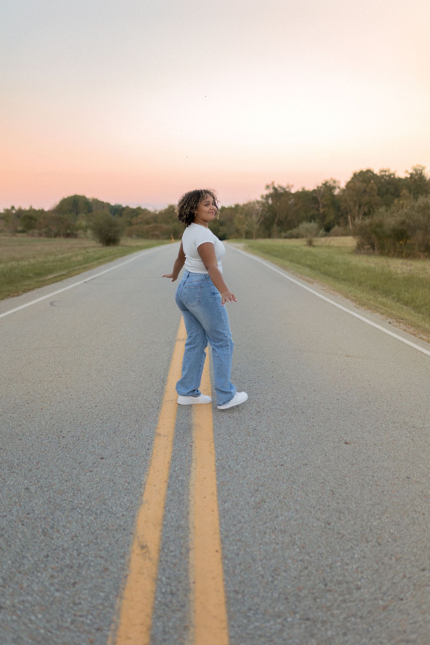 Person in casual wear standing on empty rural road during sunset.