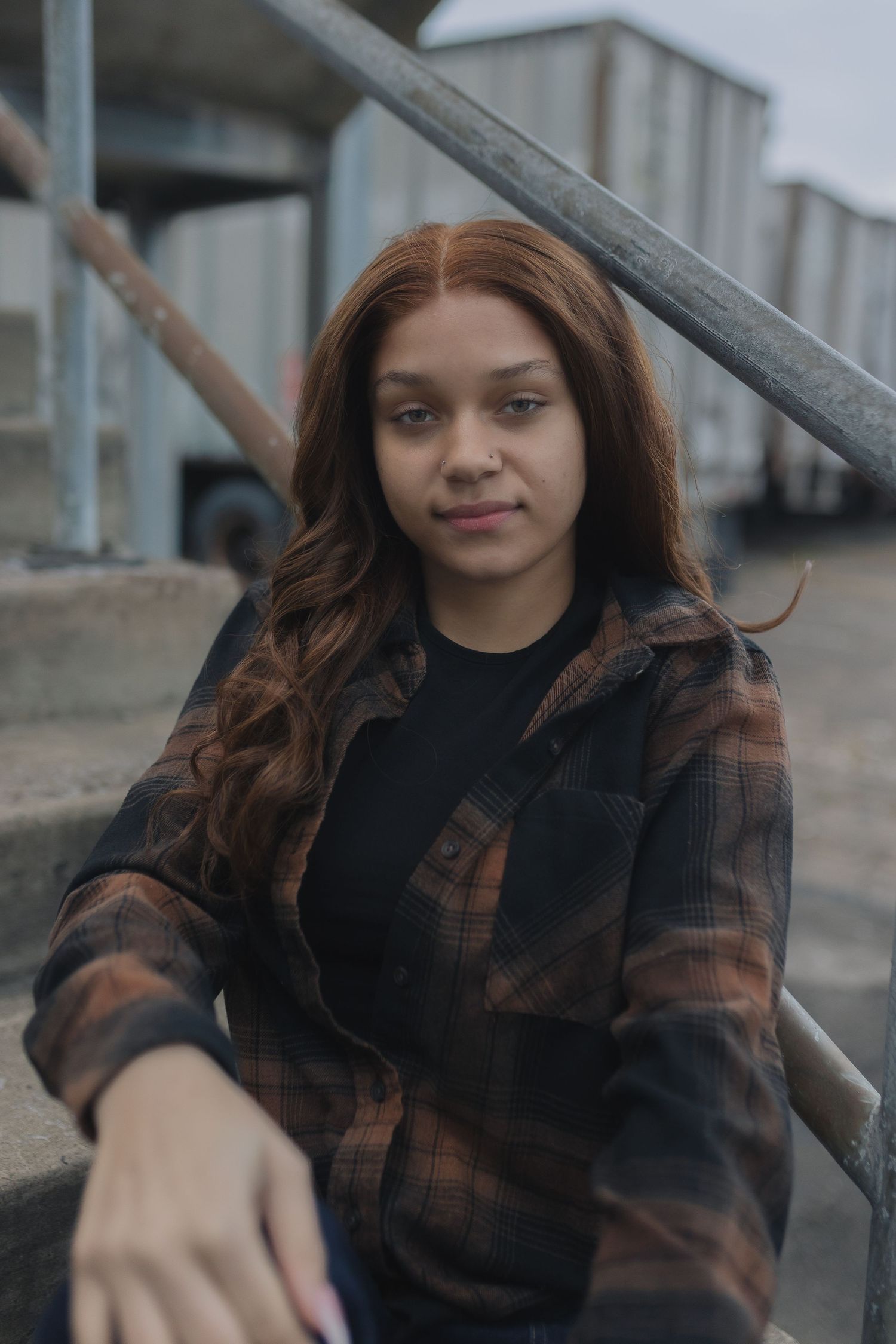 Person in brown plaid jacket poses against industrial backdrop with long wavy hair.