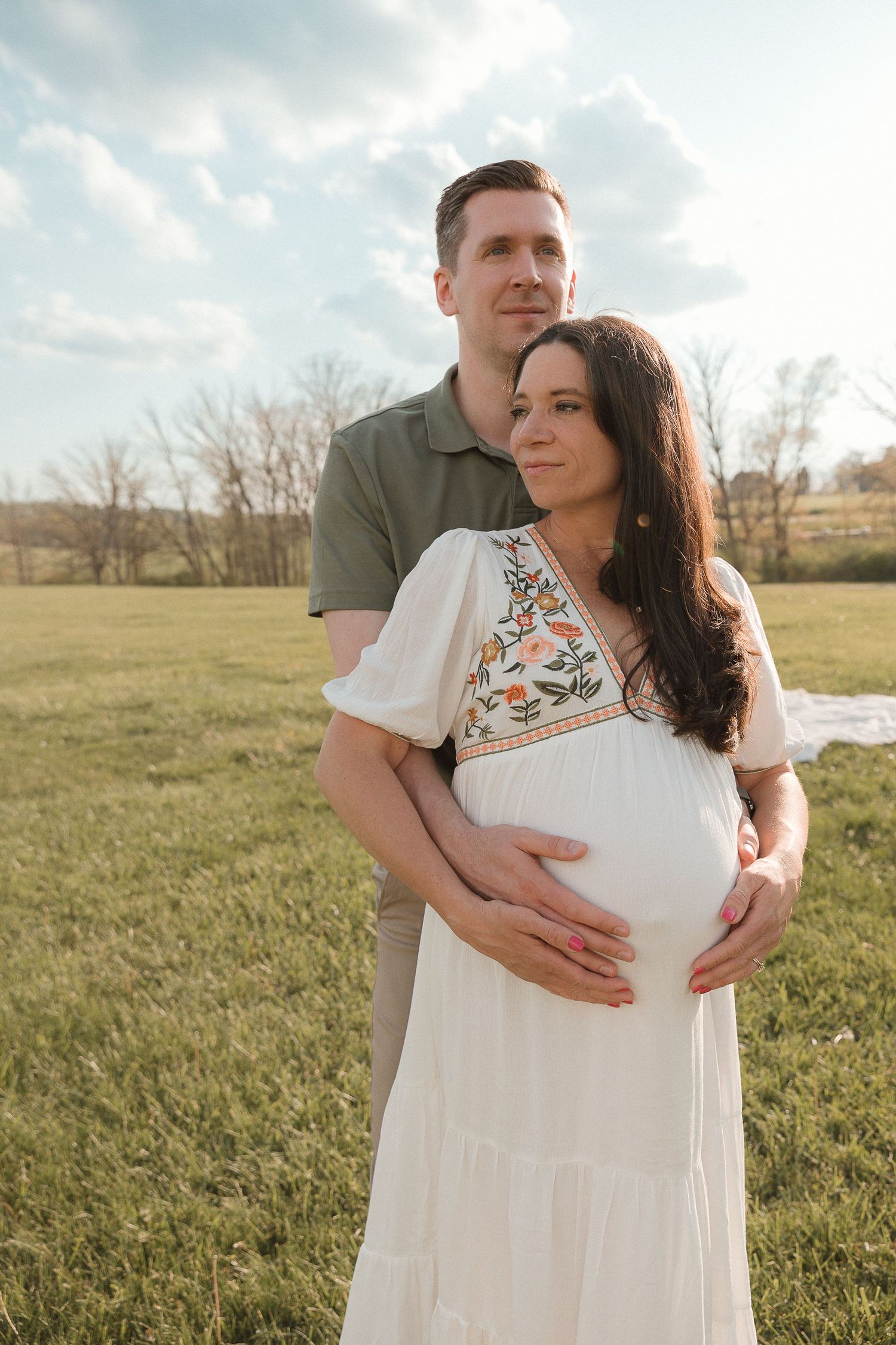 Tender outdoor maternity photo with film tones, couple embracing baby bump under soft skies with storytelling emotion.
