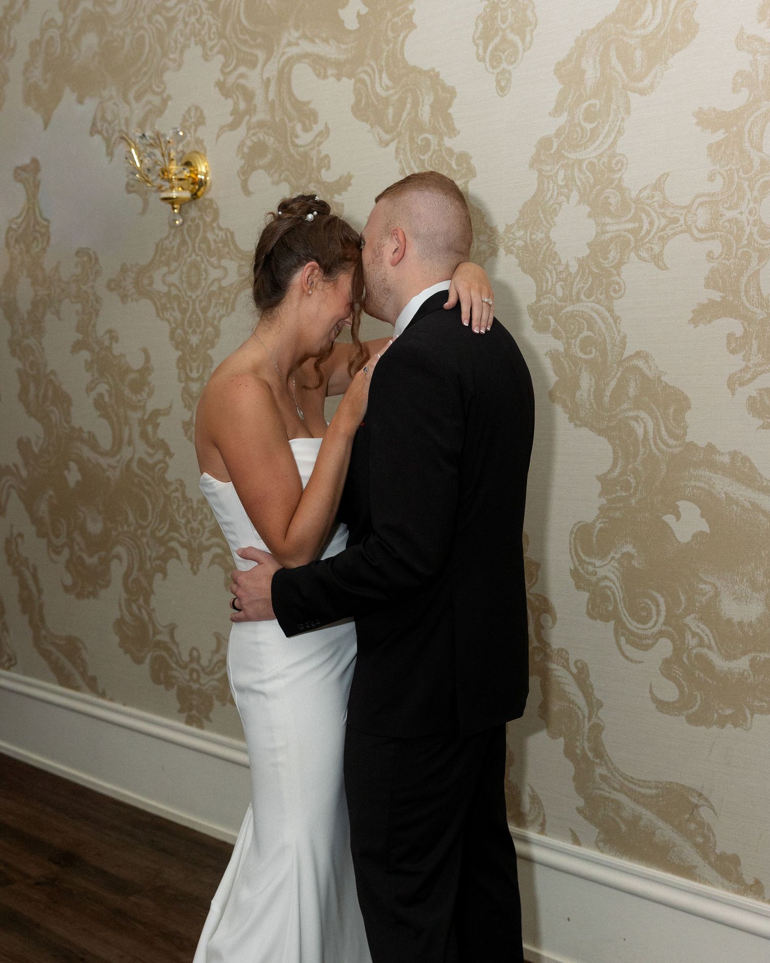 A couple in wedding attire share an intimate dance moment against a gold damask wallpaper background.