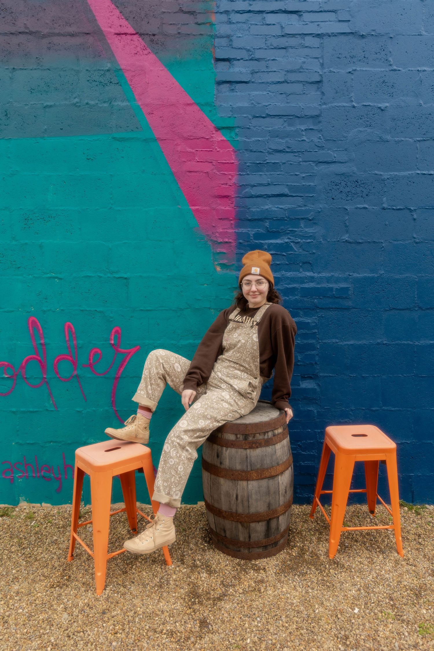 Casual street style with Carhartt beanie and overalls sitting on wooden barrel against colorful graffiti wall.