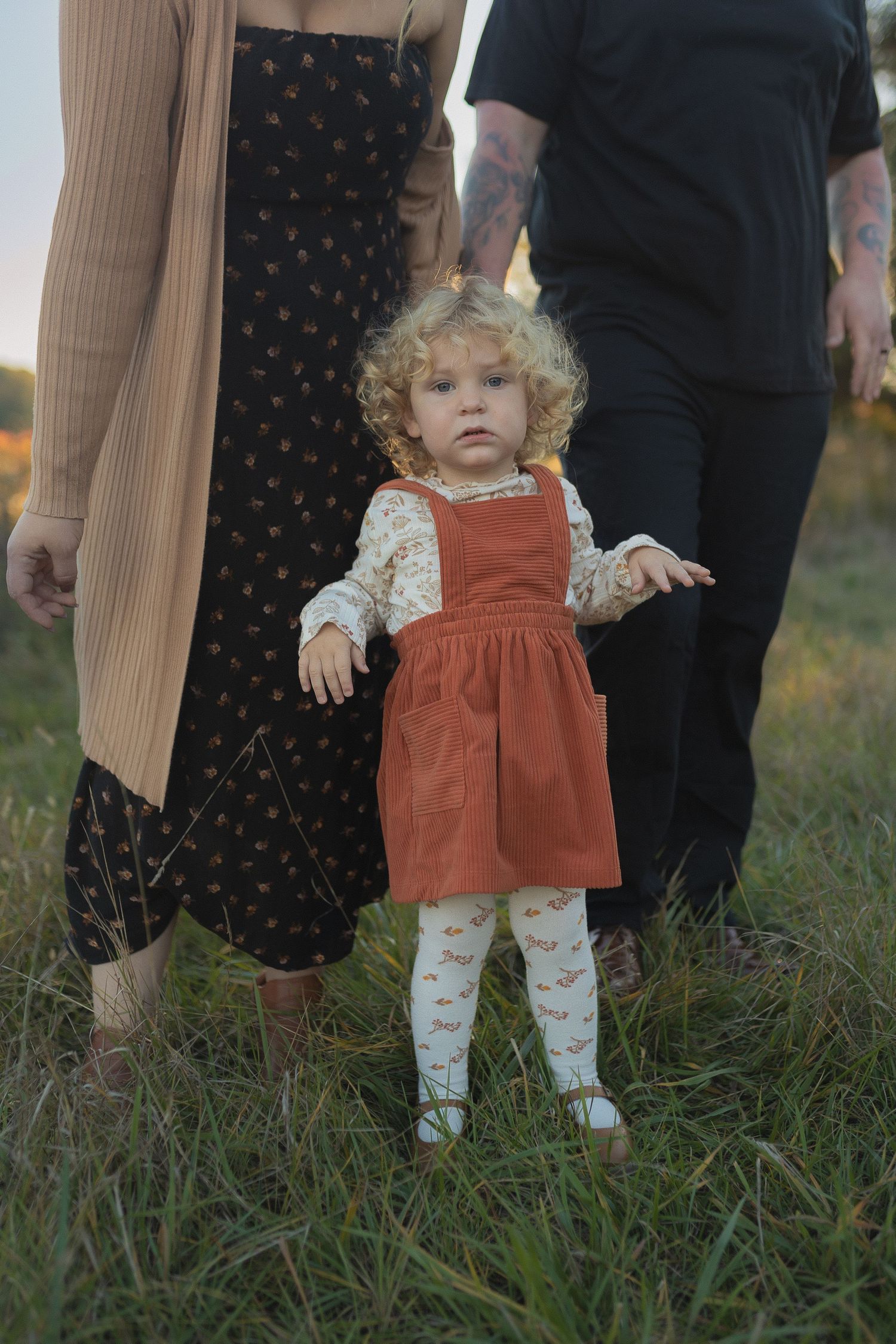 Toddler in orange pinafore dress and floral tights stands in meadow at sunset.
