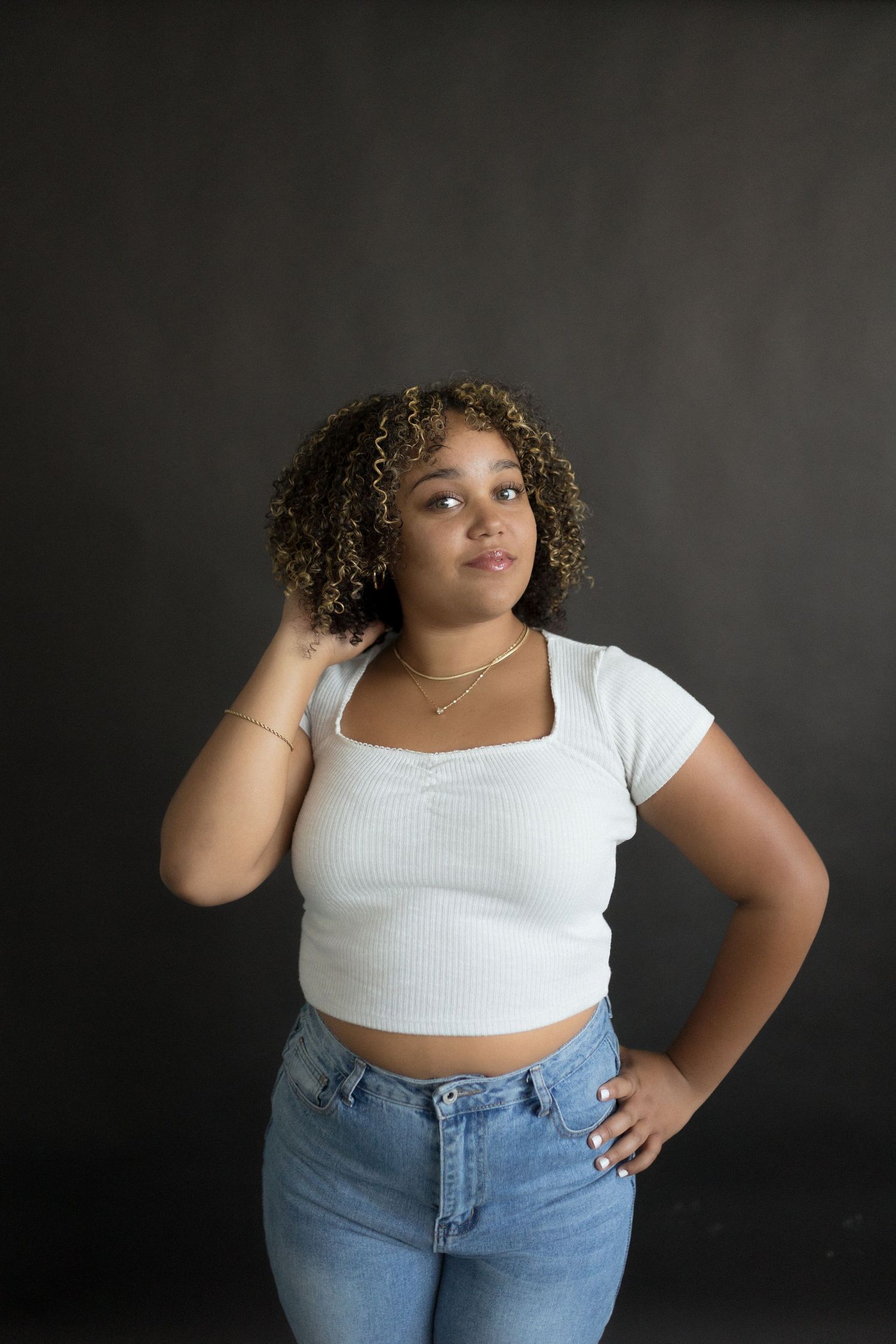 Person in white crop top and jeans posing against dark background in studio setting.