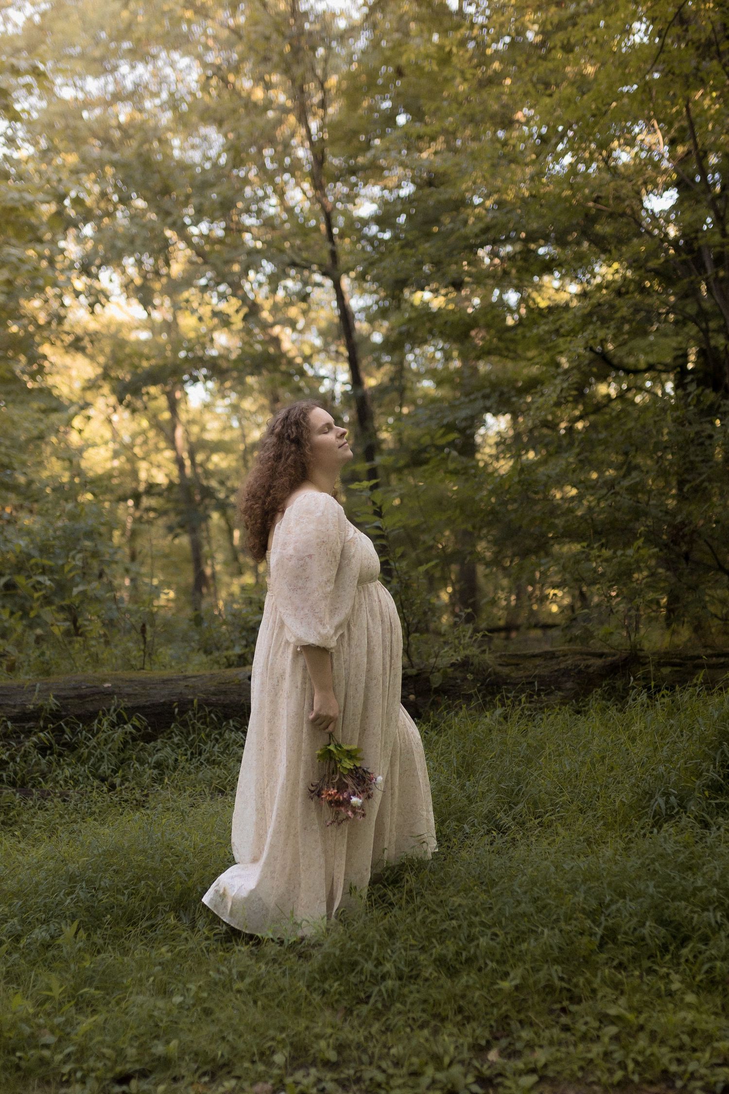Pregnant woman in white dress poses in a wooded area holding wildflowers.