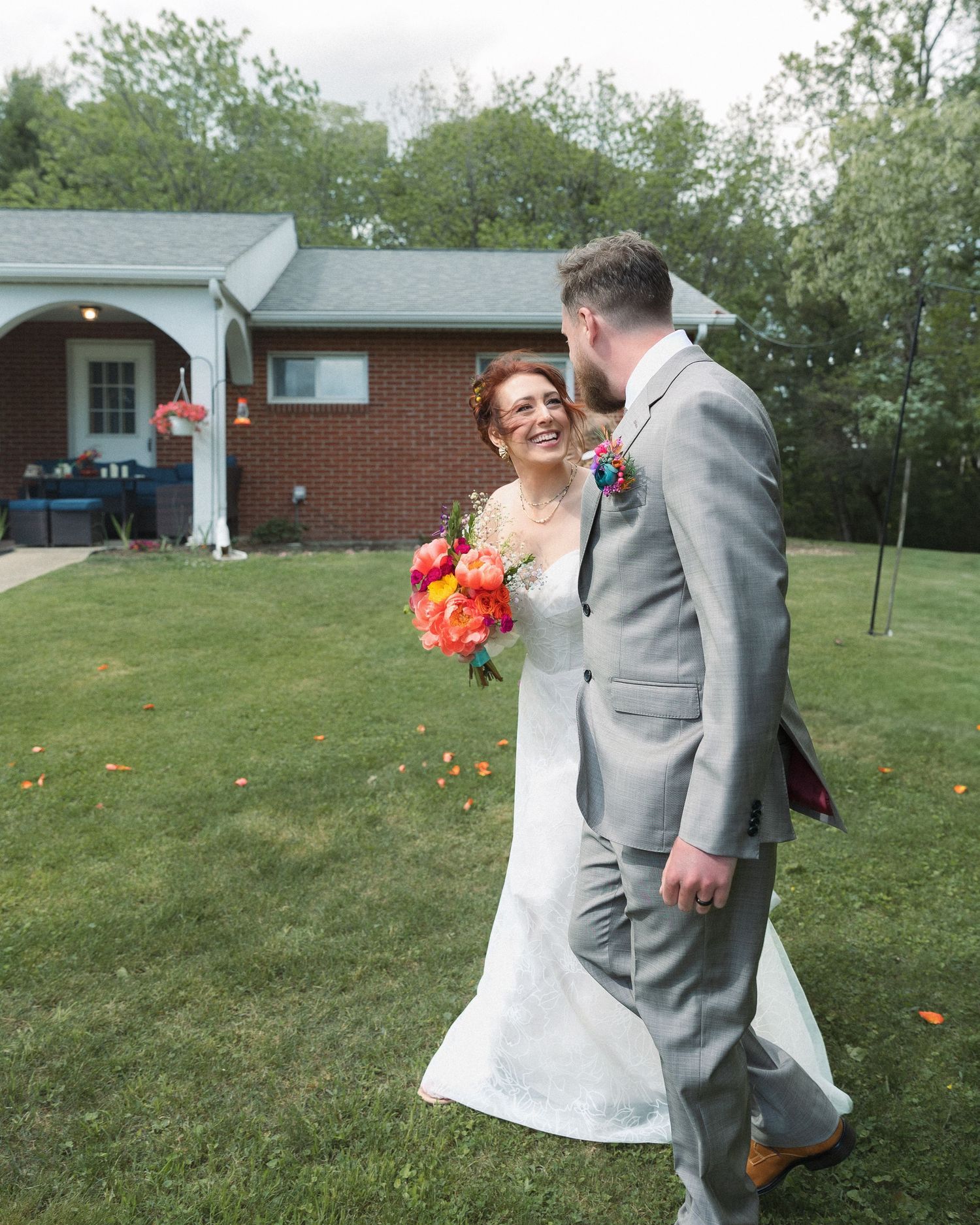 A wedding couple walks together outside on a green lawn holding vibrant red and orange bouquets.