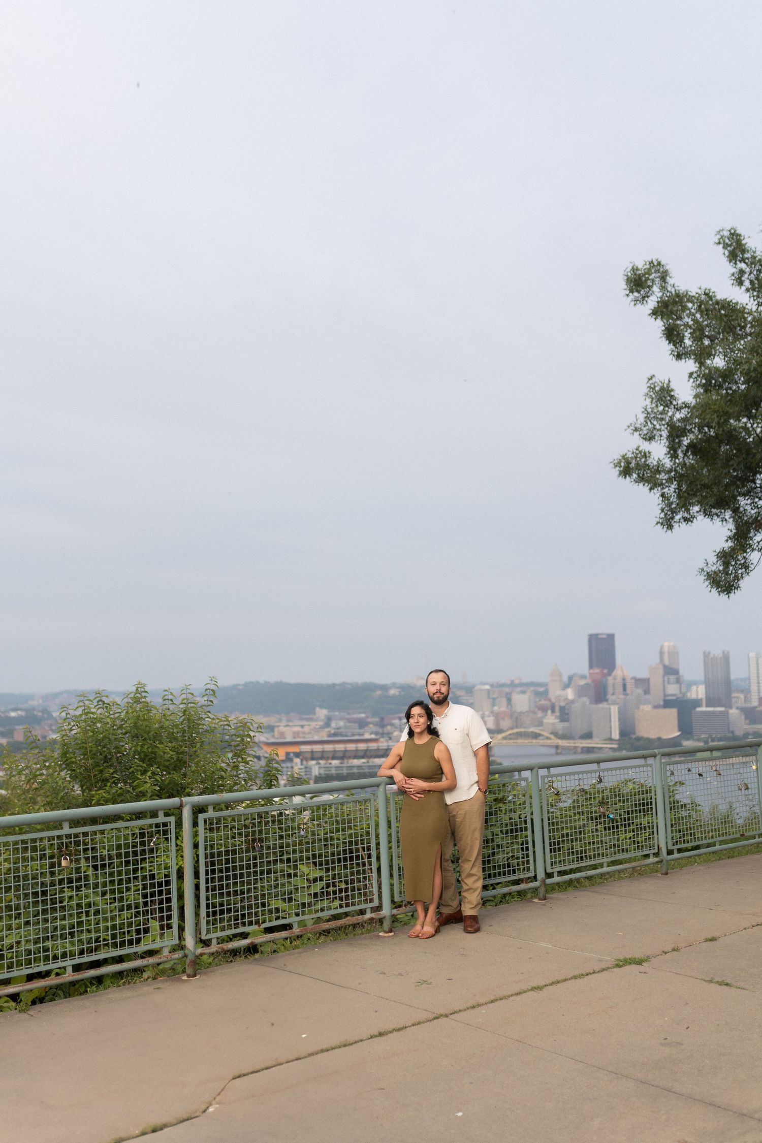 A couple standing at scenic overlook with Pittsburgh skyline visible in background.