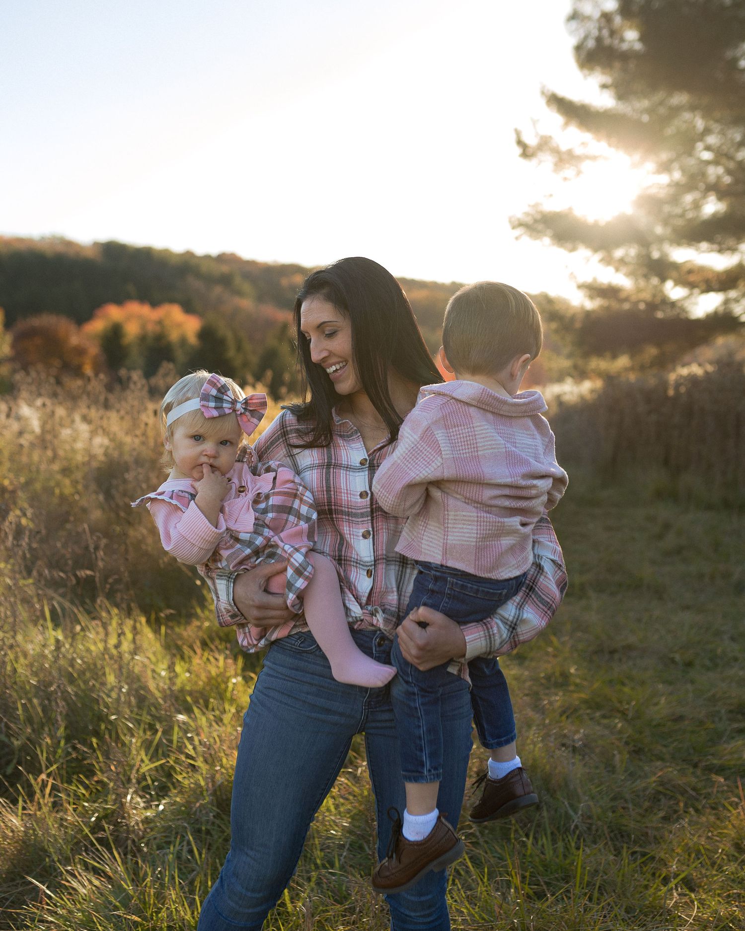 Woman holding two small children in casual wear during golden hour in a field.