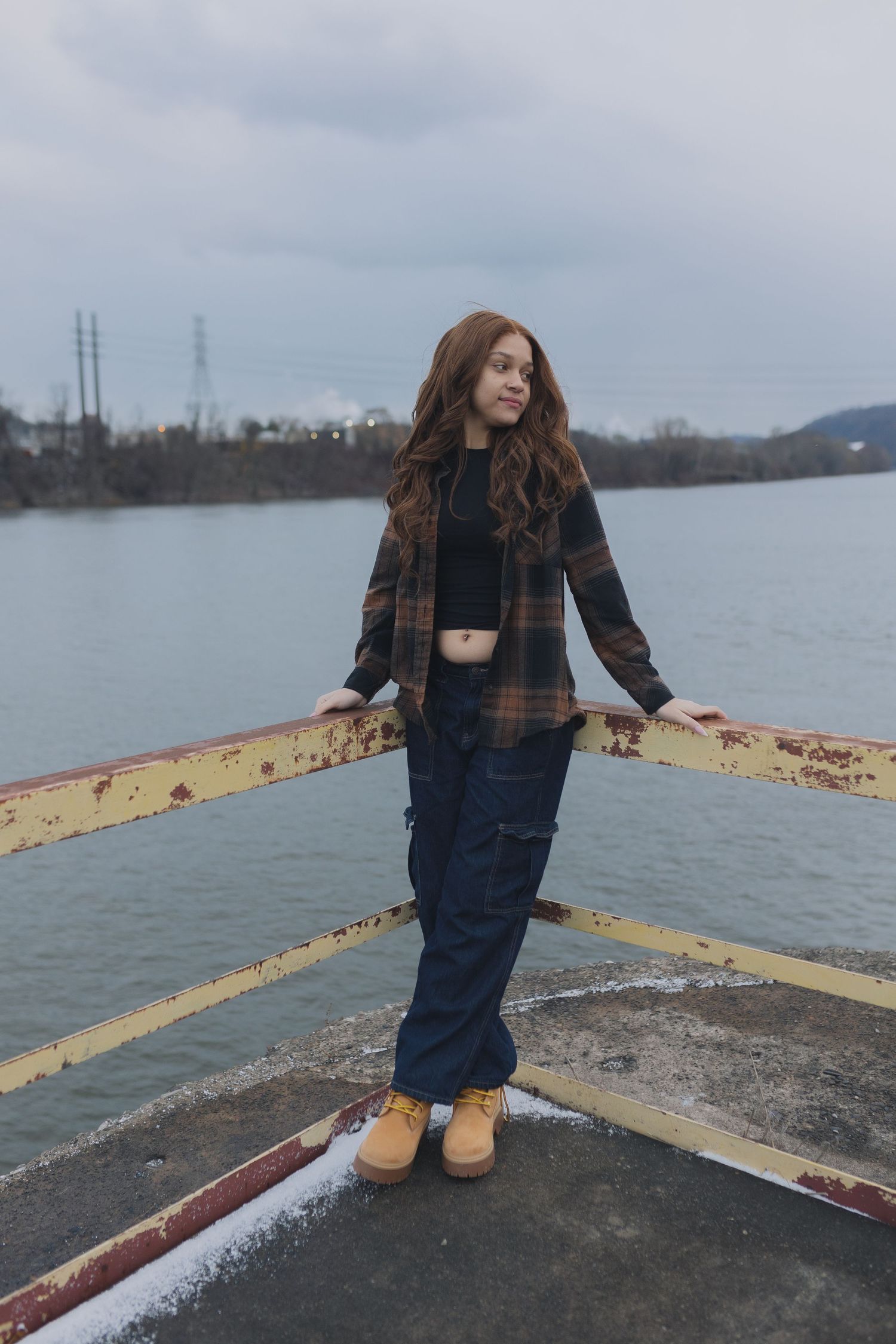 Person in plaid jacket and jeans stands on concrete pier overlooking lake on cloudy day.