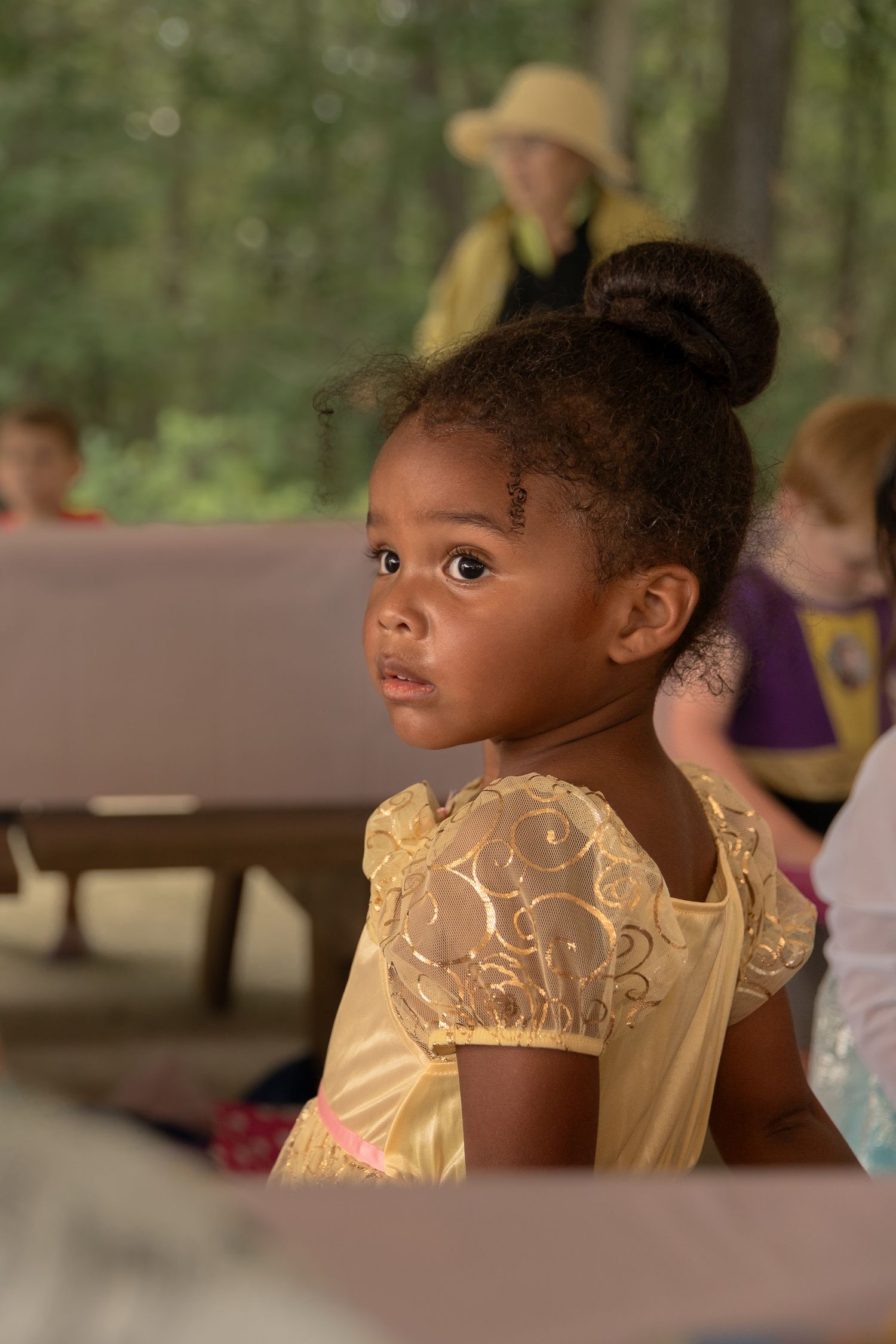 A child in a beautiful yellow princess dress sits thoughtfully at an outdoor gathering.