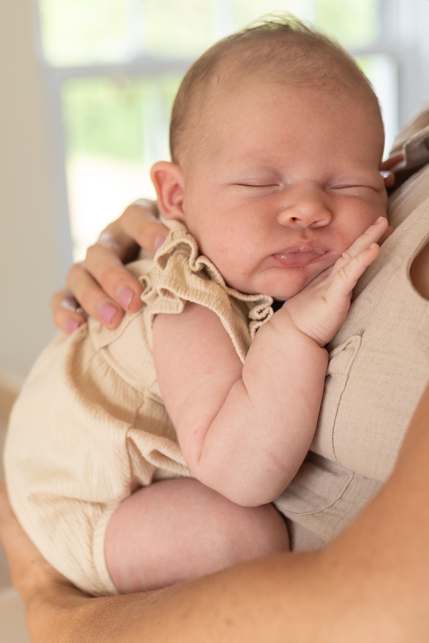 A sleeping infant in a beige outfit rests peacefully against someone's shoulder.