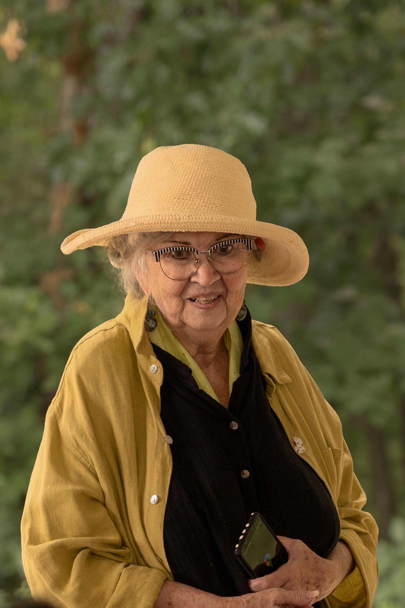 An elderly person wearing a straw hat and yellow jacket smiles warmly in a natural setting.