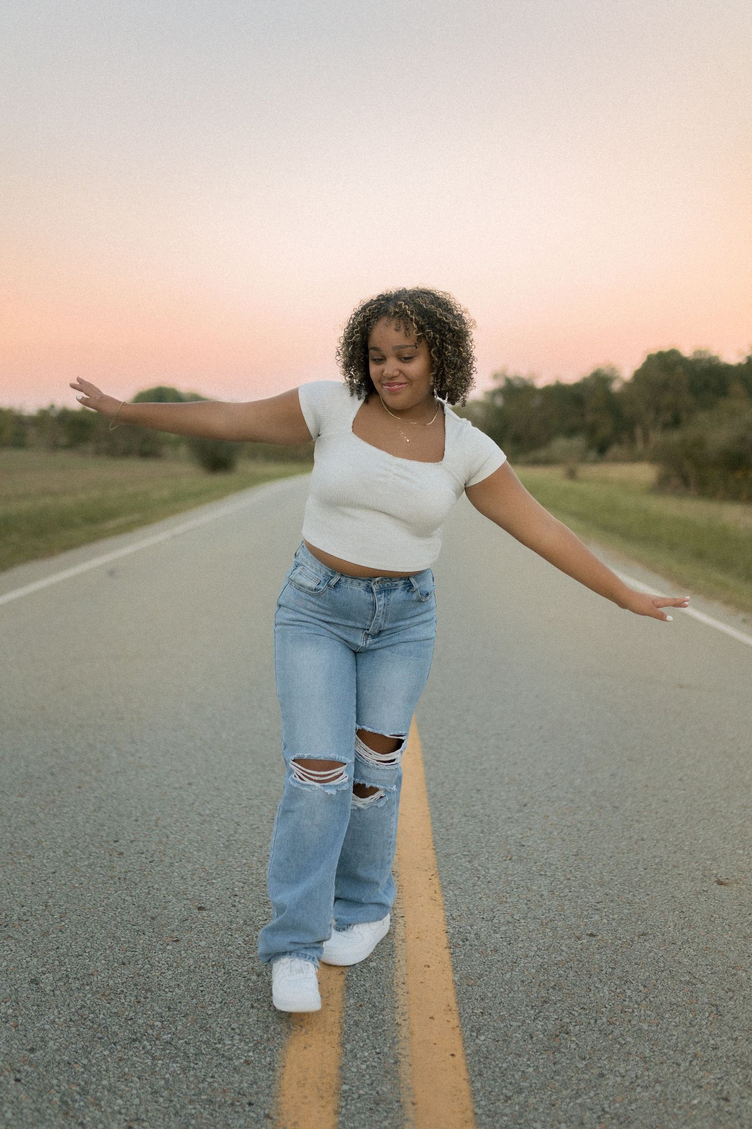 Person balancing on yellow road line wearing ripped jeans and white crop top at sunset.