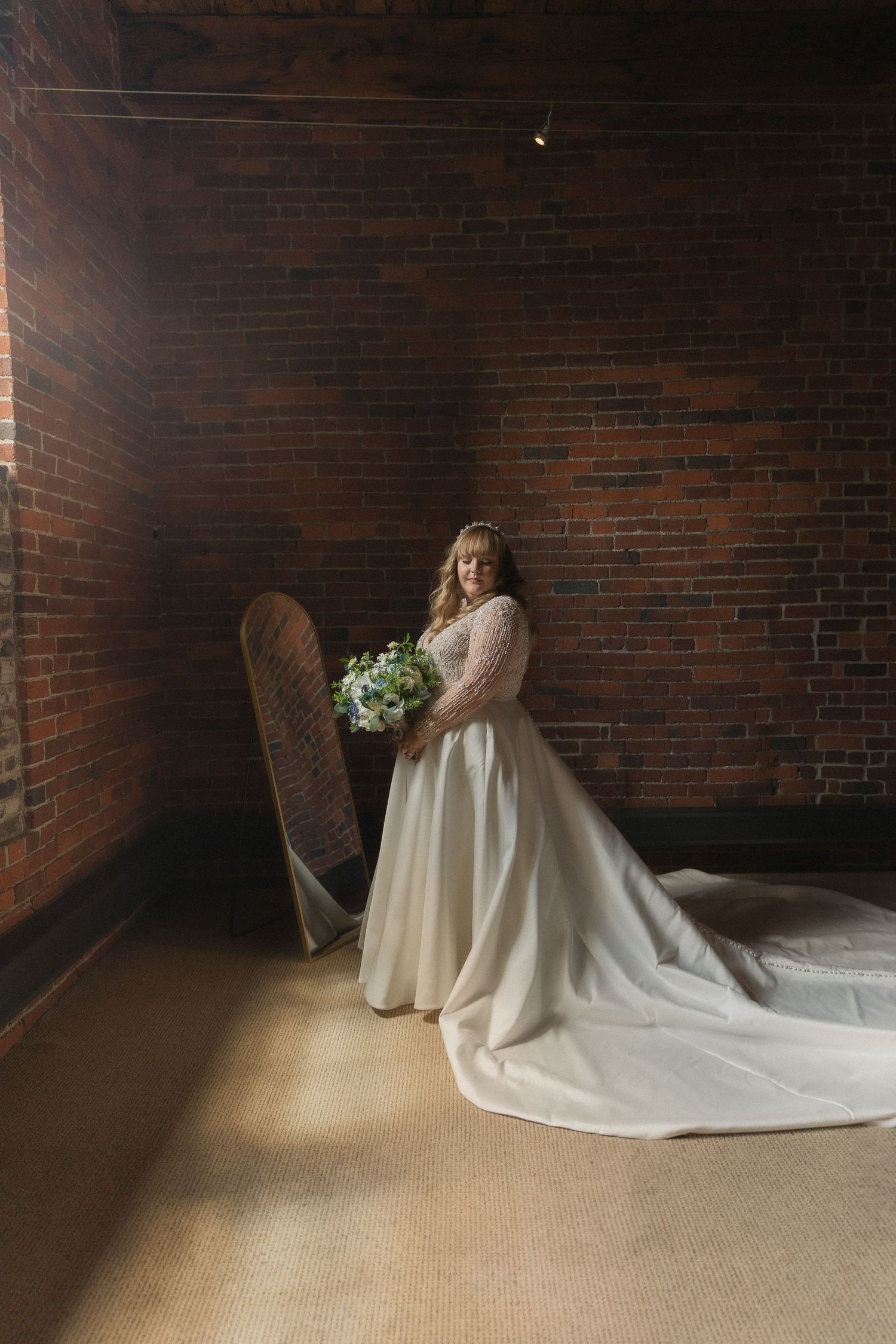 Bride in elegant white wedding gown poses against rustic brick wall with bouquet.