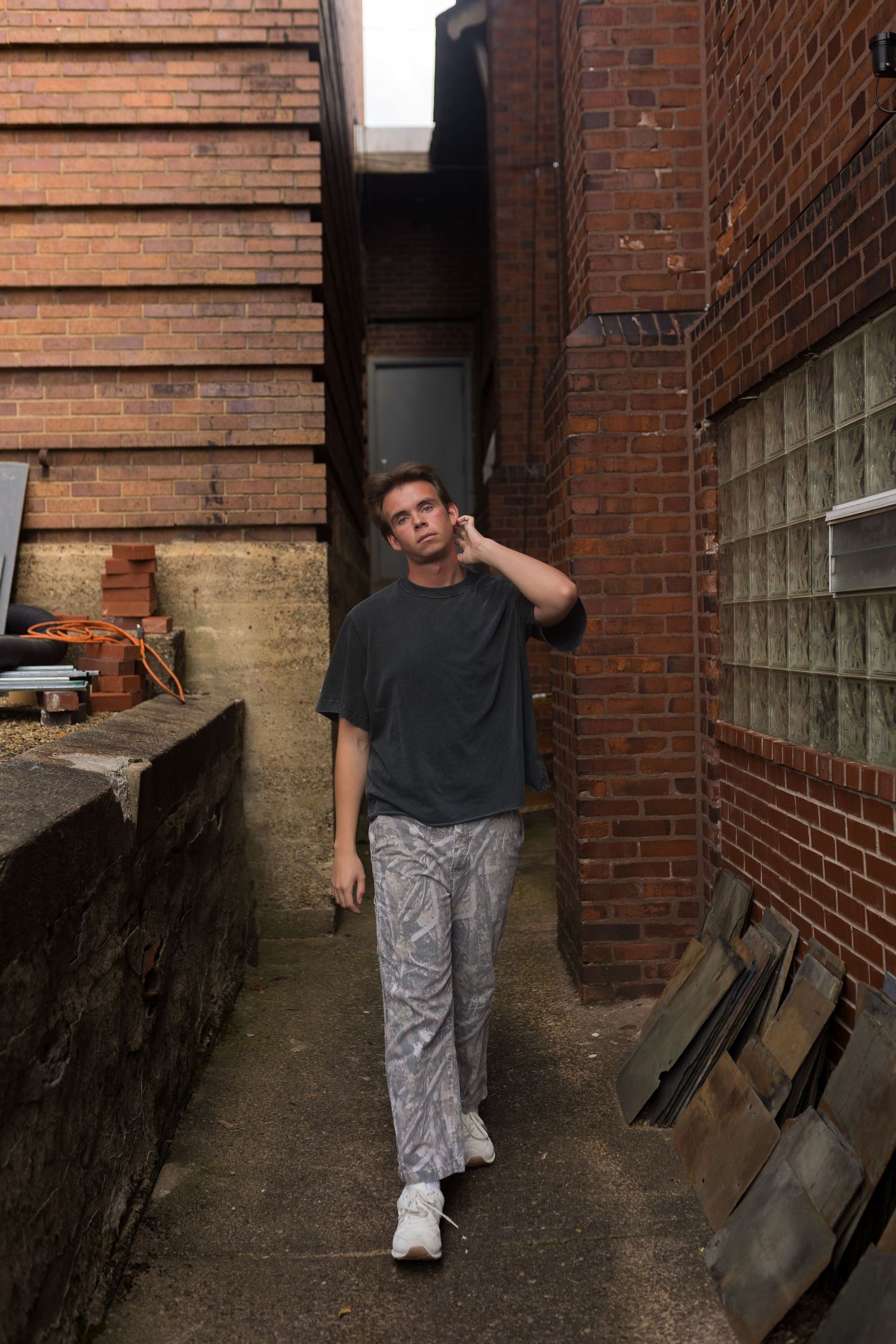 Person walks through narrow brick alleyway wearing dark t-shirt and patterned pants.