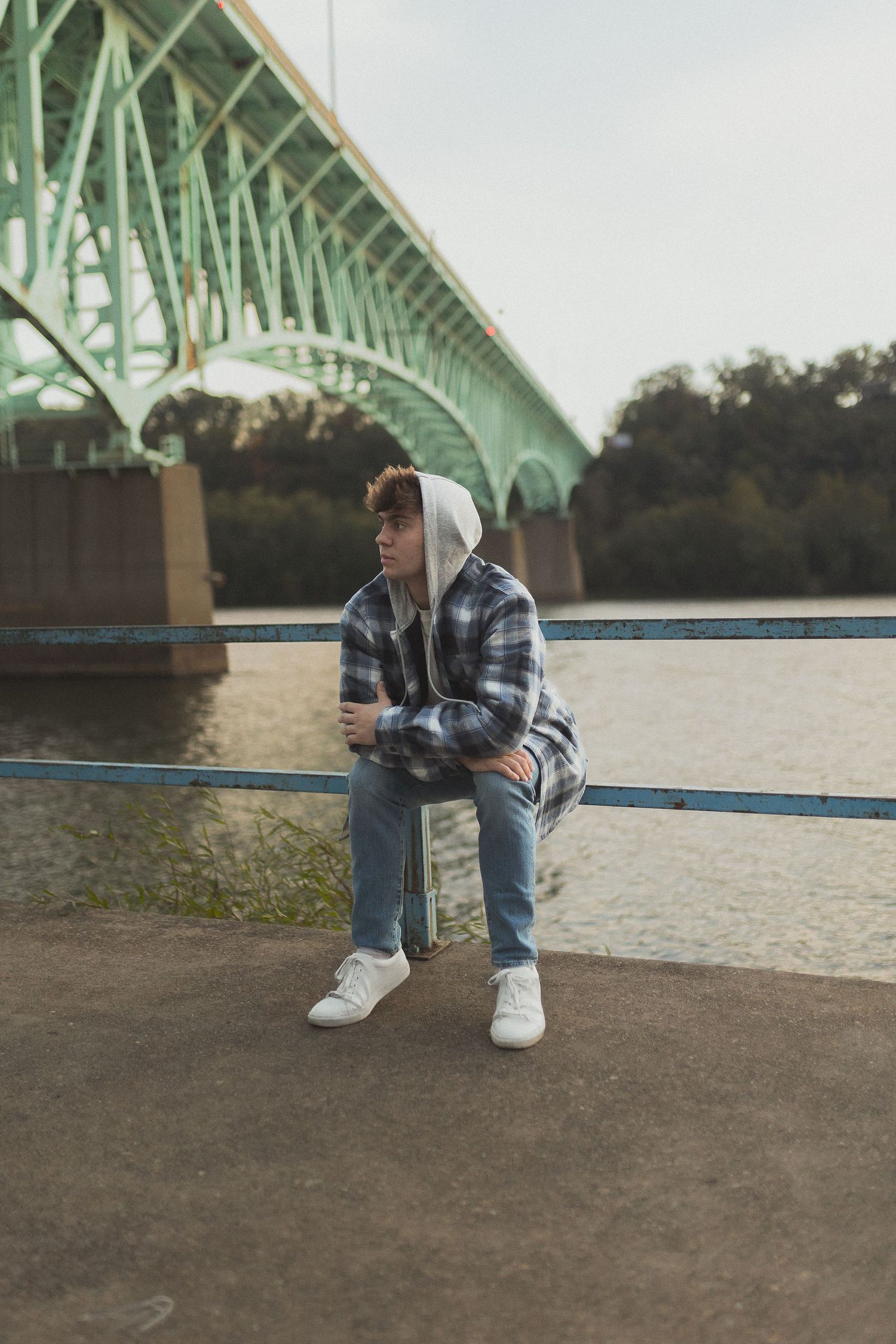 Person in plaid jacket sitting on railing with large green bridge in background.