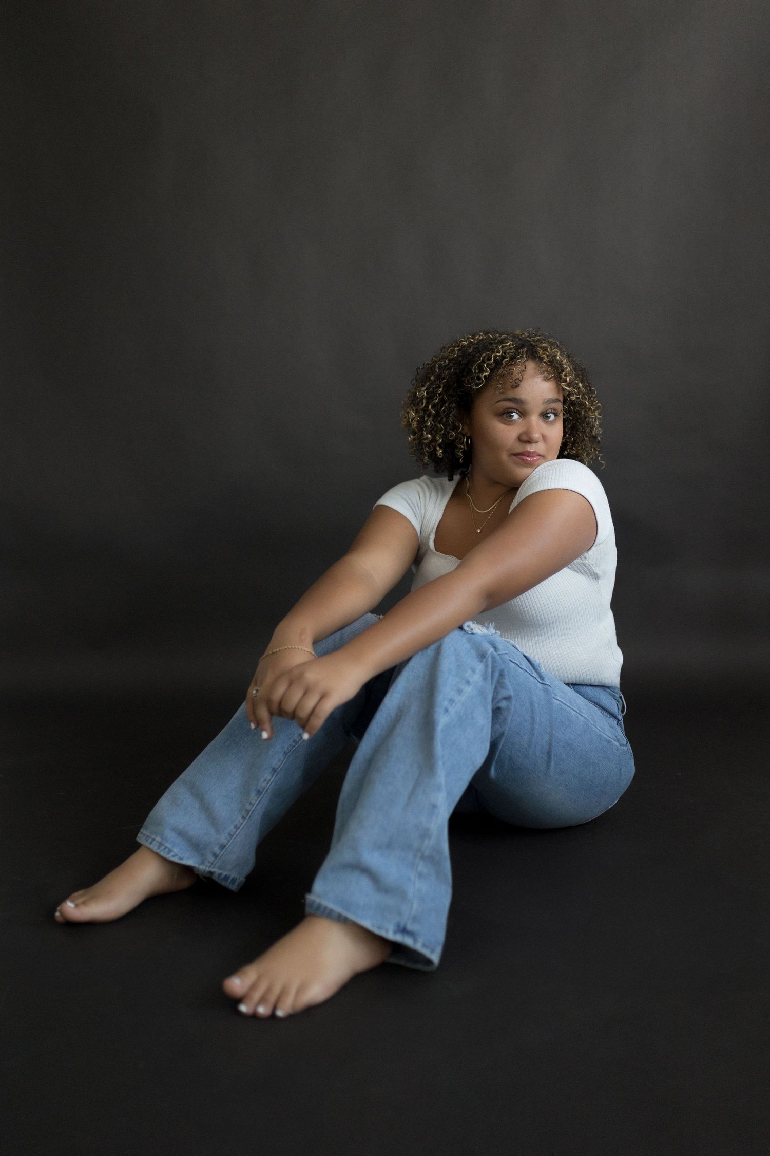 Person in white tank top and blue jeans sitting barefoot against dark background.
