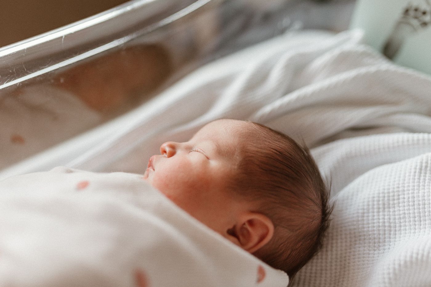 Close-up profile view of newborn baby sleeping peacefully in bassinet, wrapped in soft white swaddle with peachy dots, golden hour light.
