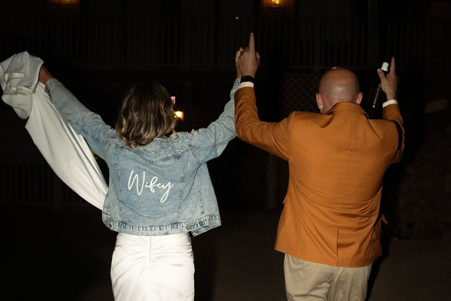 A couple celebrating at night with raised arms, bride wearing customized denim jacket.