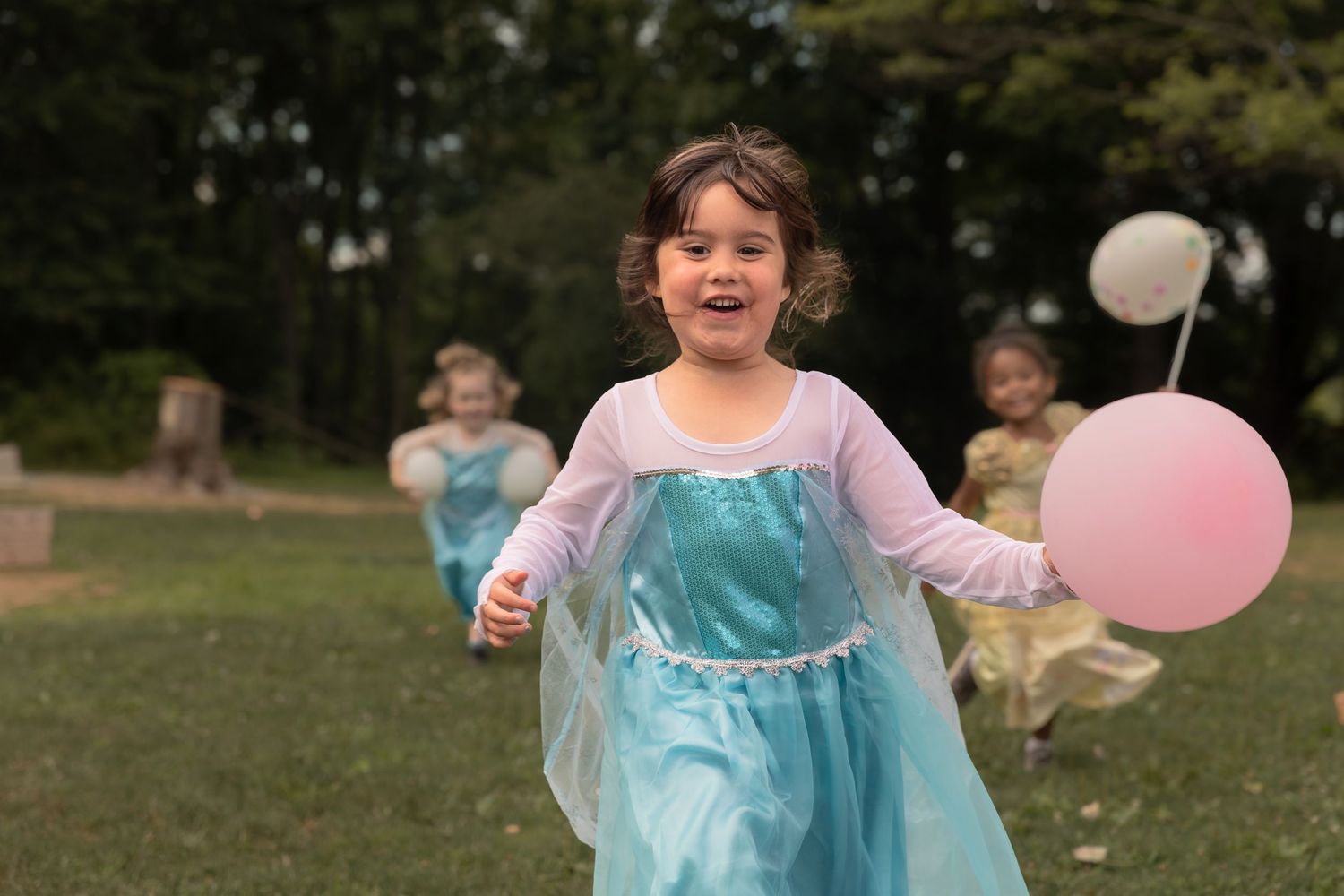 A joyful child running outdoors in a blue princess dress with pink balloons in the background.