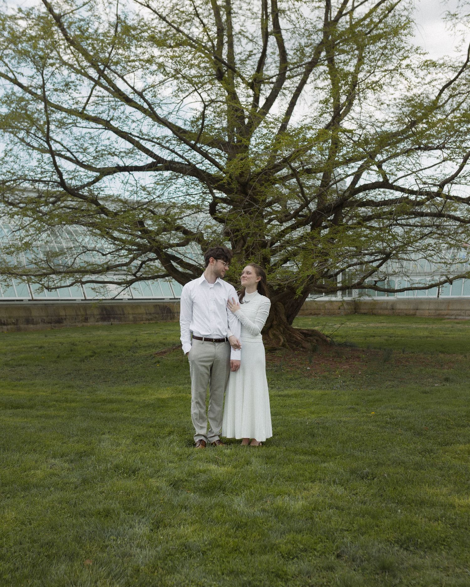 A couple in formal attire stands hand-in-hand on green grass beneath a large tree on a cloudy spring day.