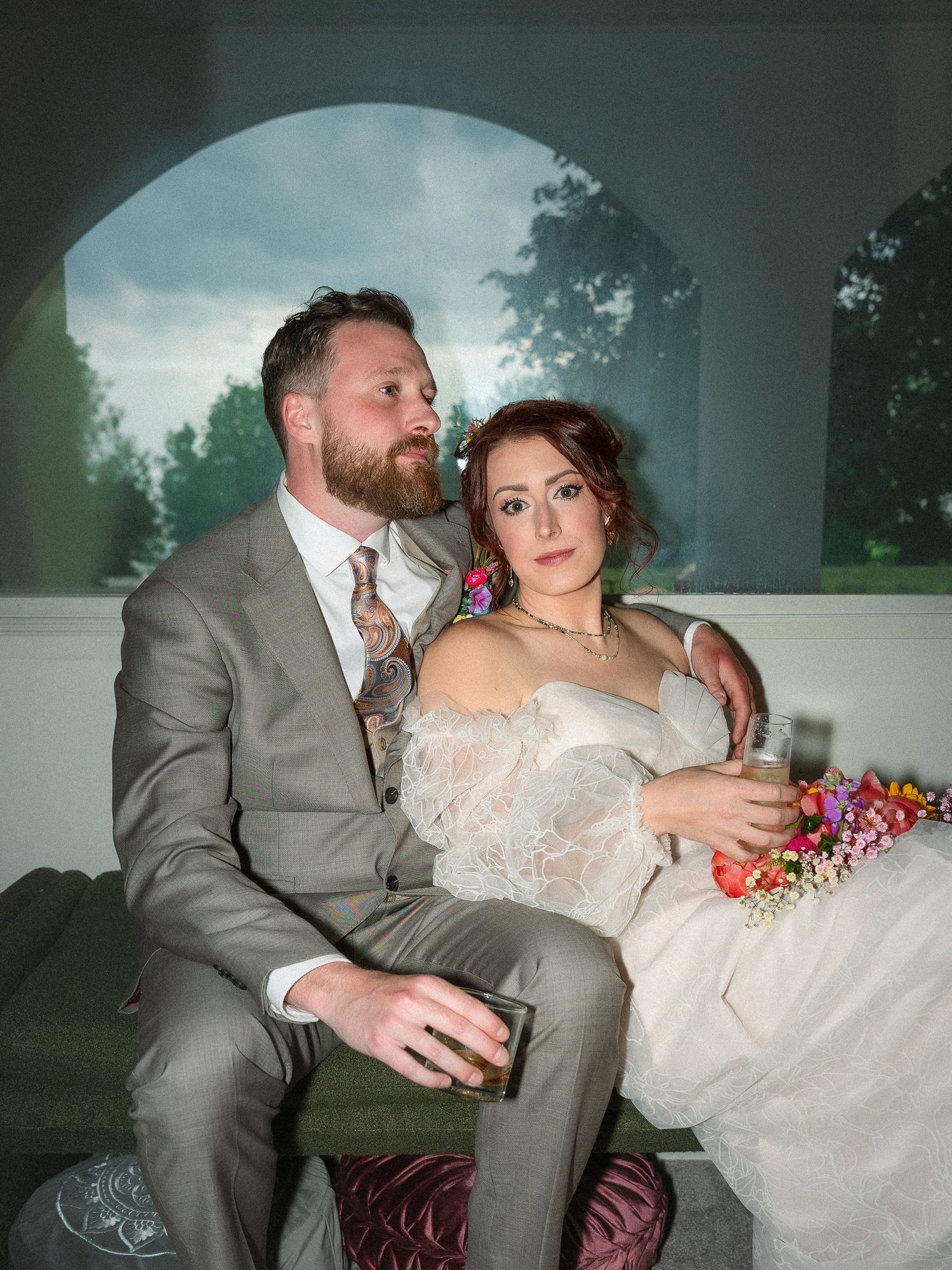 A wedding couple playfully poses on a teal velvet couch during their outdoor reception at dusk.