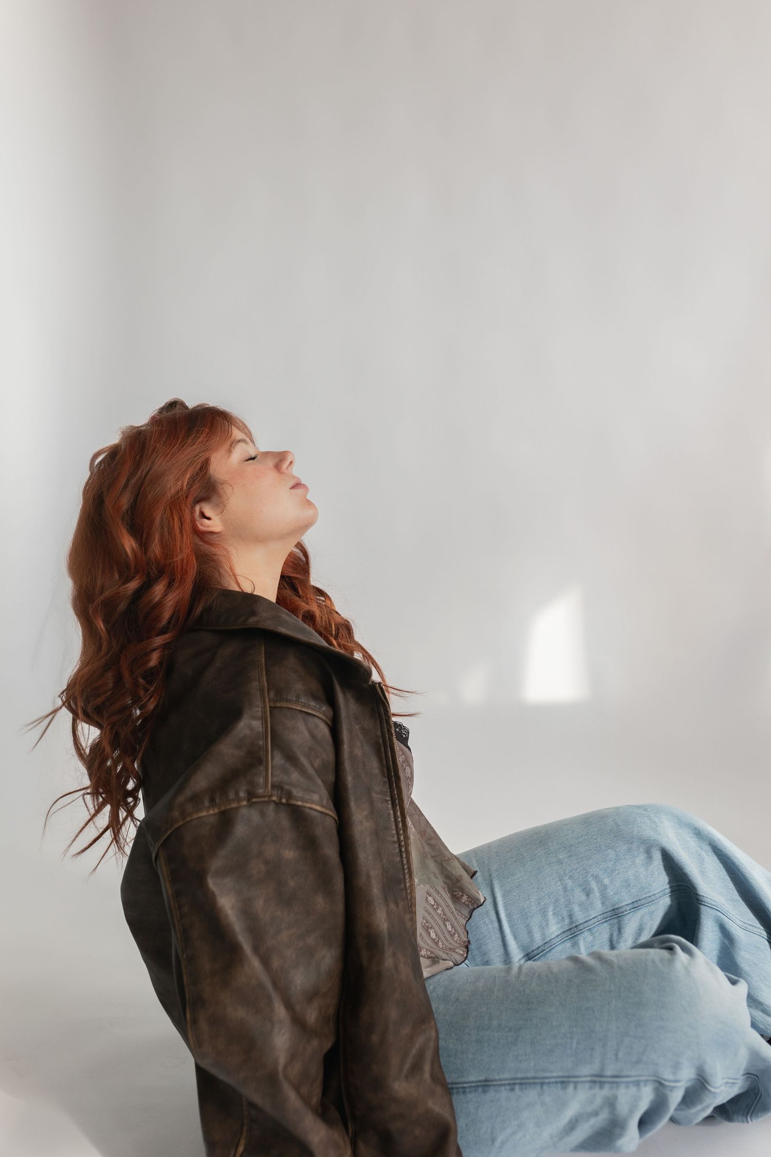 Person wearing brown leather jacket and jeans poses against white wall with dramatic lighting.