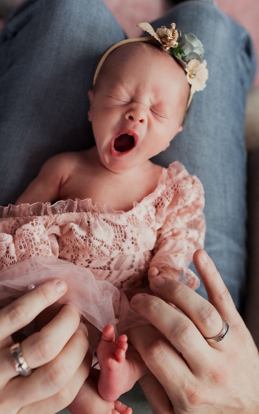 Newborn baby girl in blush dress and floral headband, captured mid-yawn embraced by parent's hands during a sweet in-home lifestyle session.