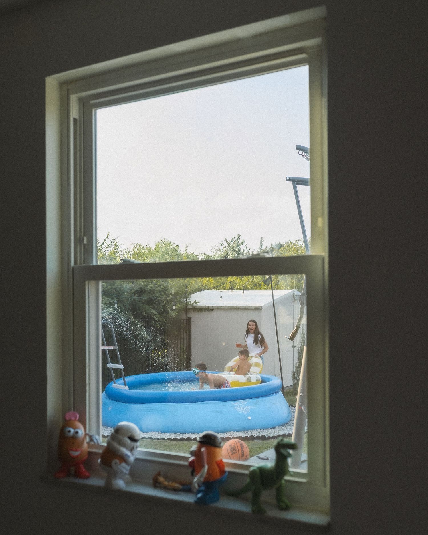View through a window showing a blue inflatable pool in a backyard with toy figures lined up on the windowsill.
