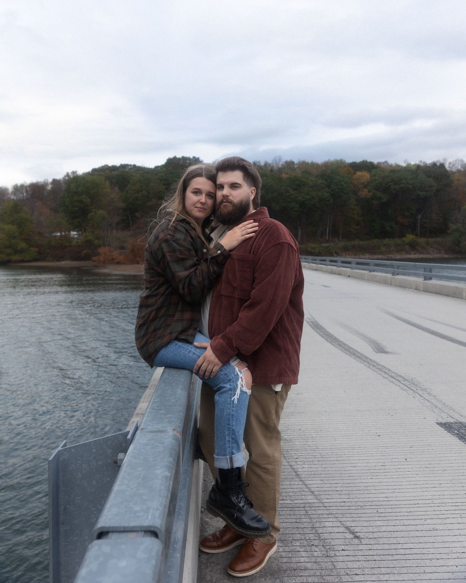 A couple embraces on a concrete bridge over a lake during an overcast autumn day.