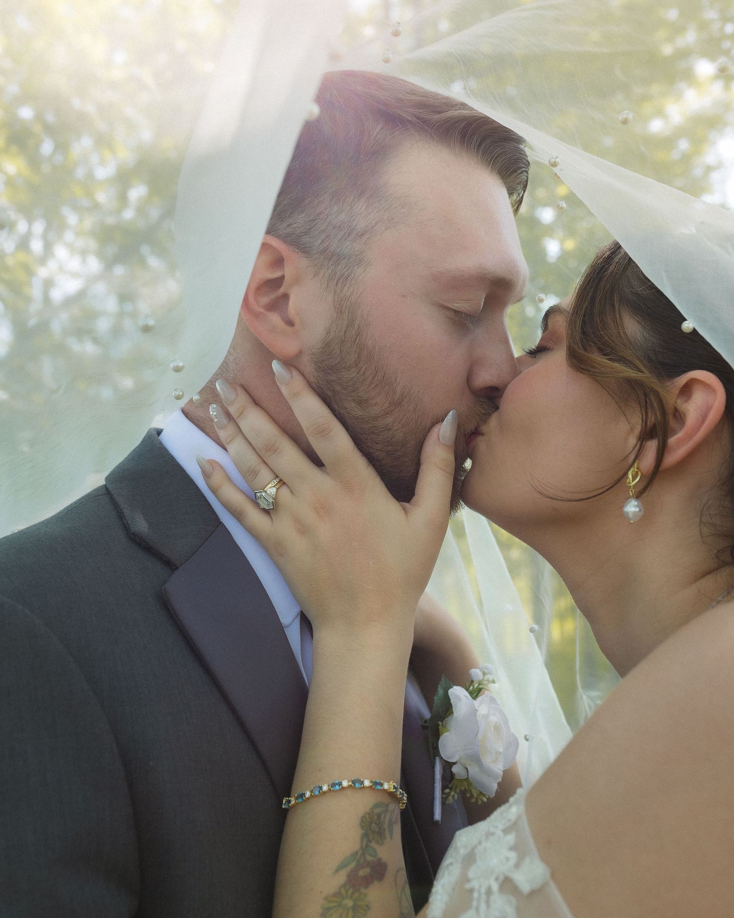 A bride and groom share a romantic kiss under a flowing veil, showcasing her diamond ring and gold bracelet.