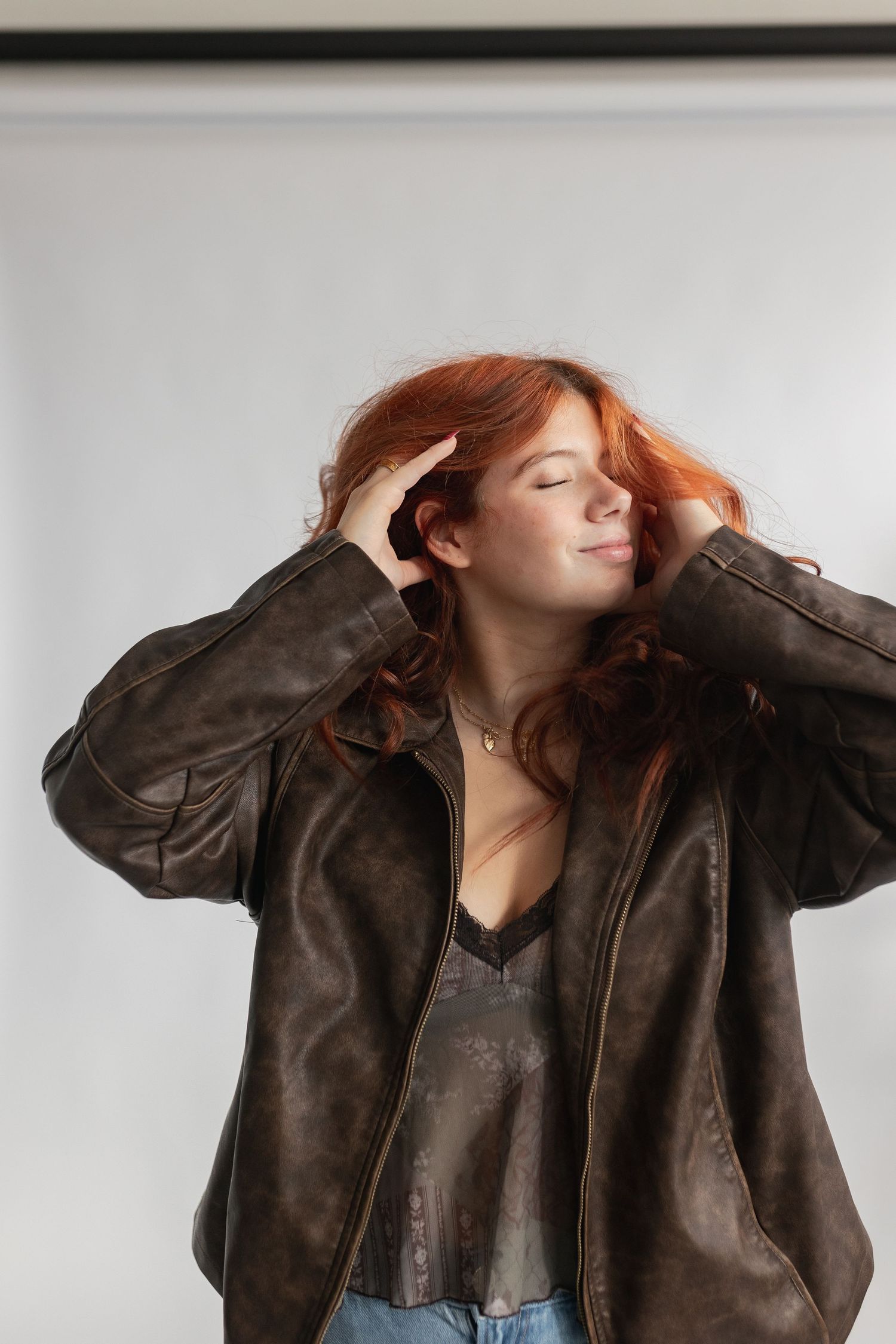 Artistic portrait in brown leather jacket with hands in hair against white background.
