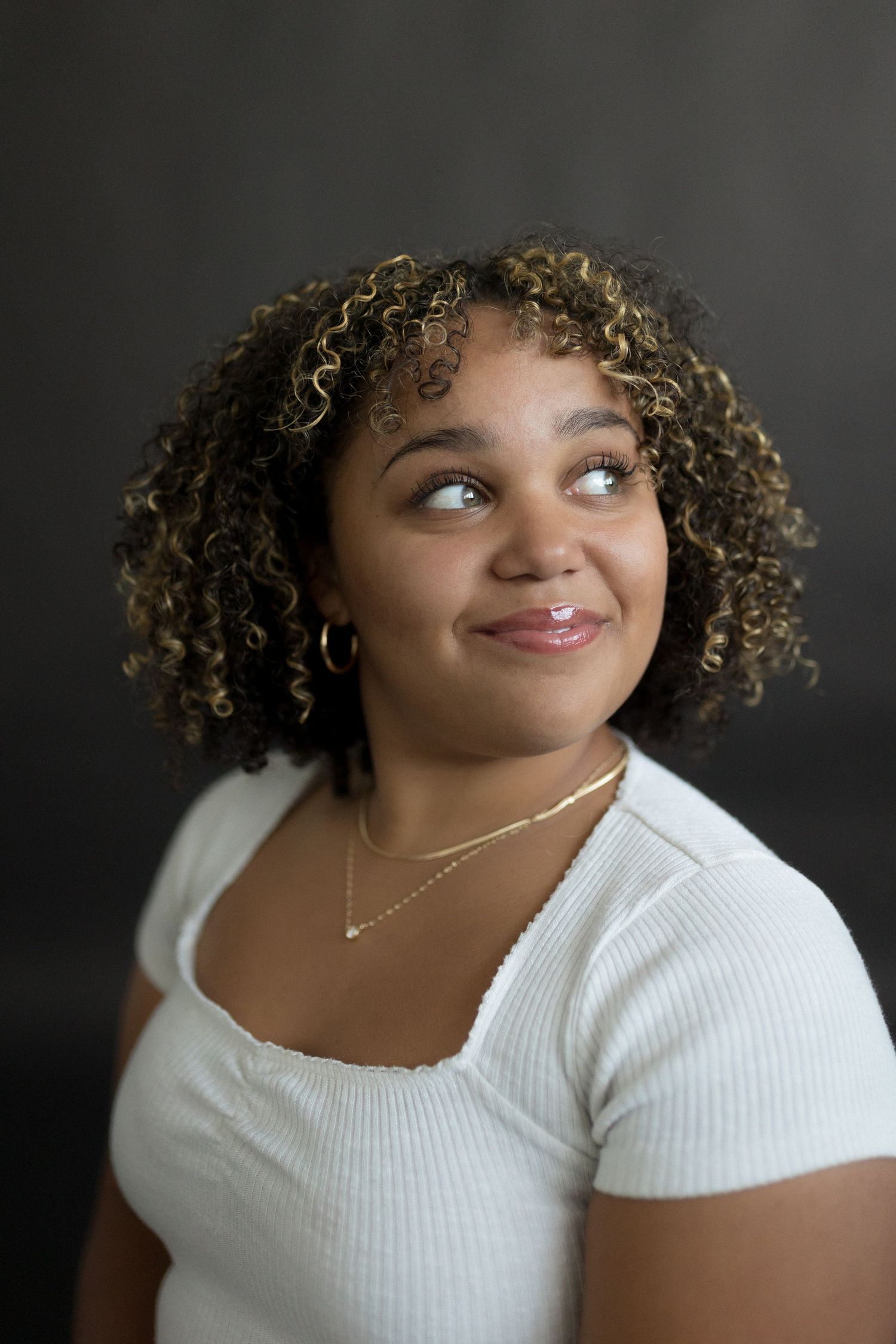 Portrait of a person in a white top with curly hair glancing sideways against a dark background.