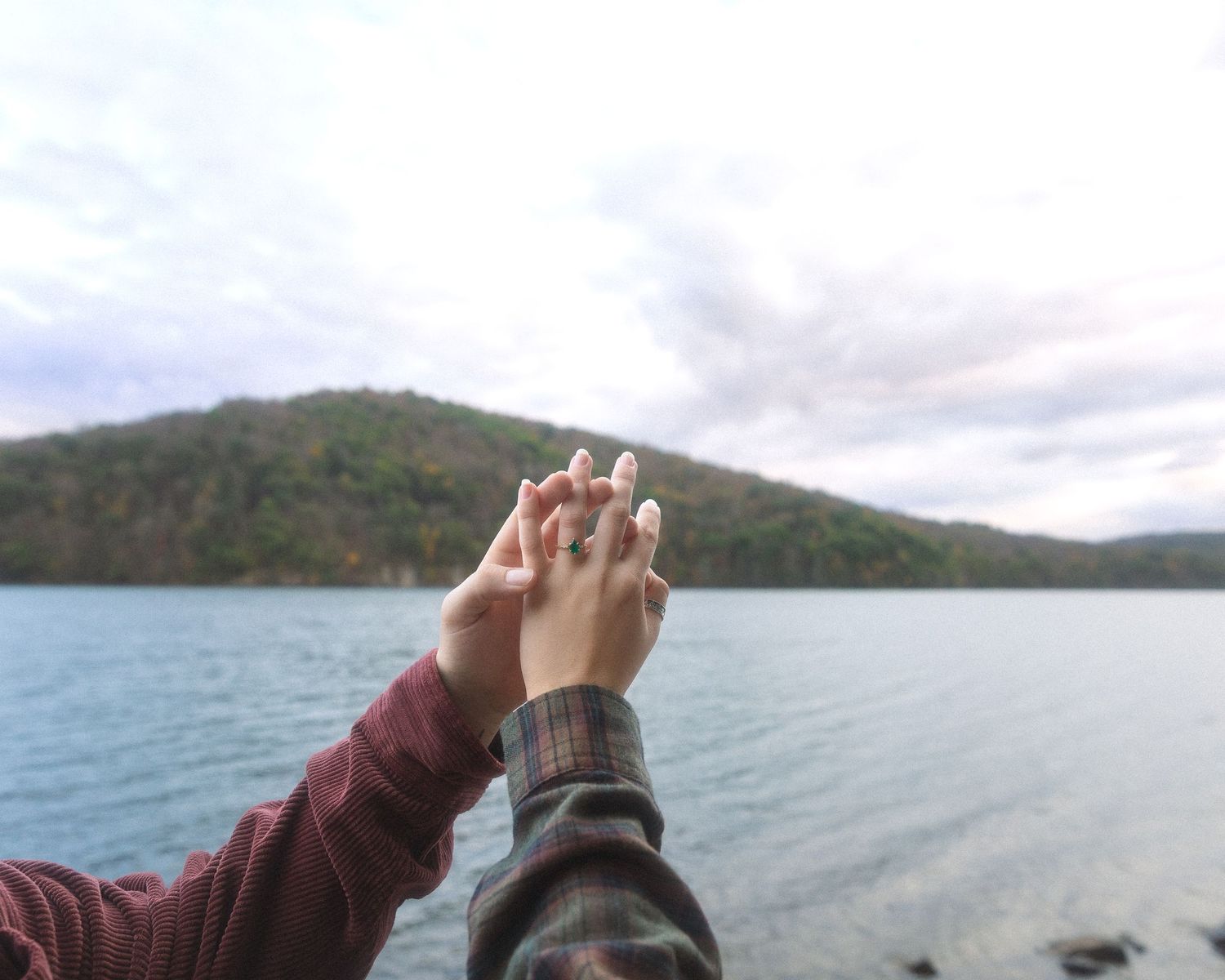 A person in a maroon sweater points towards a scenic mountain lake view on a cloudy day.