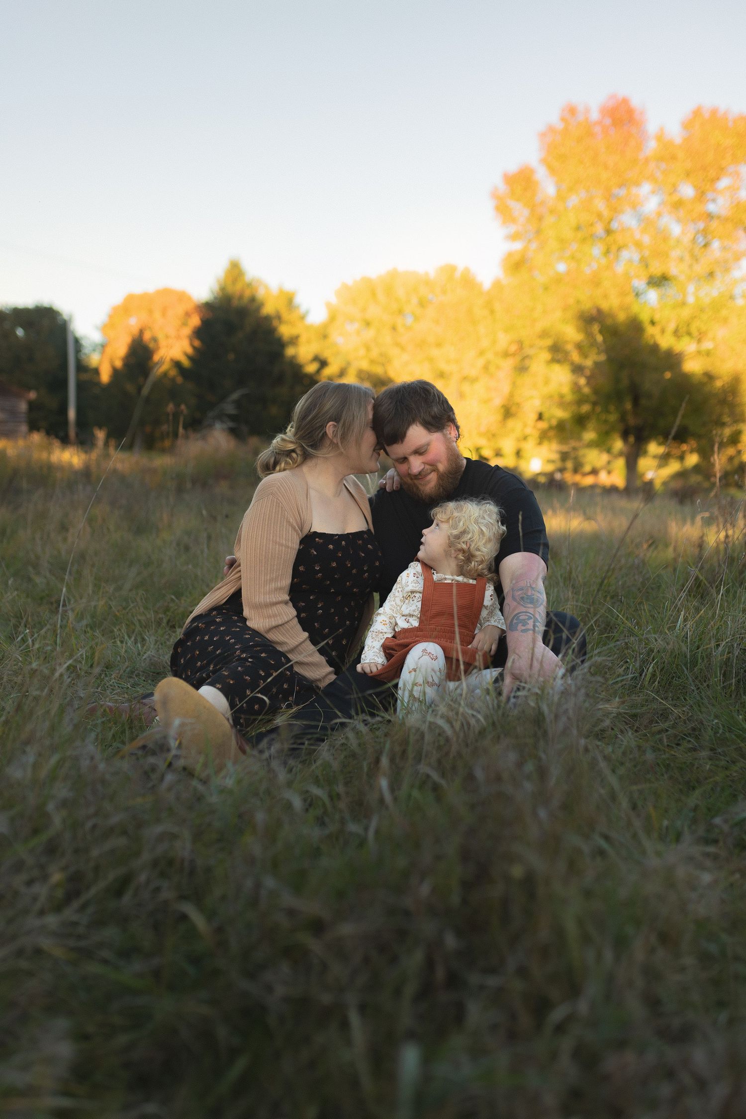 Family sits together in tall grass during golden hour sunset with autumn trees in background.