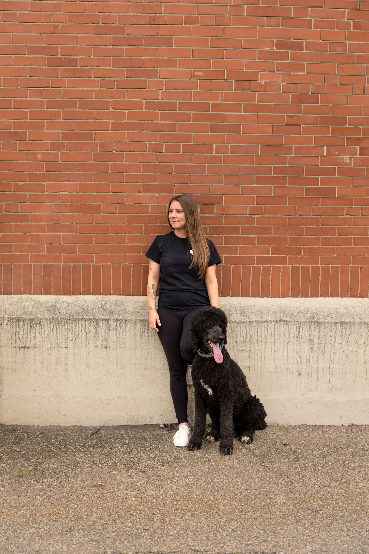 Person in black outfit standing with large black dog against brick wall.