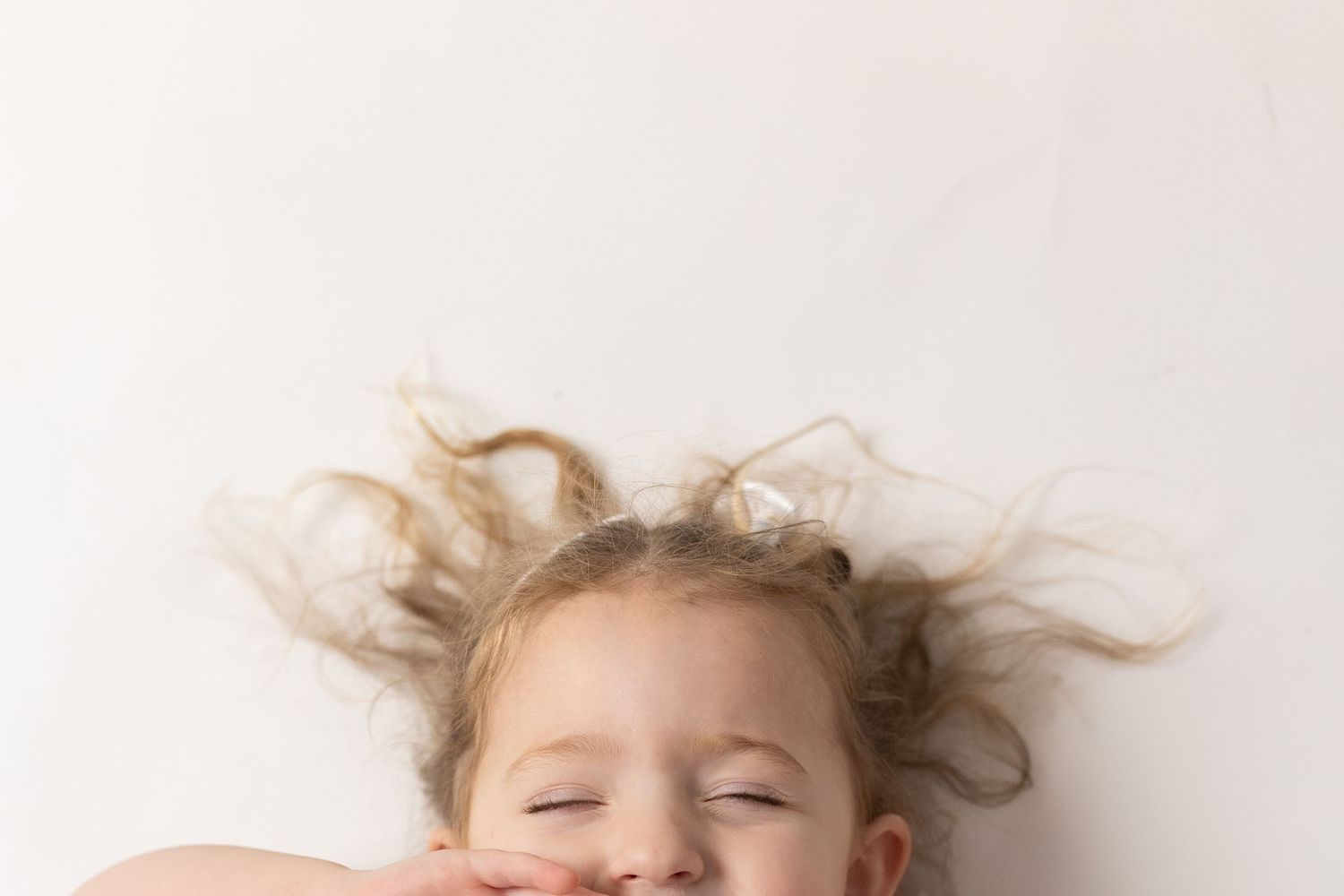 Close up of a child laying down with messy blonde hair spread out against white background.