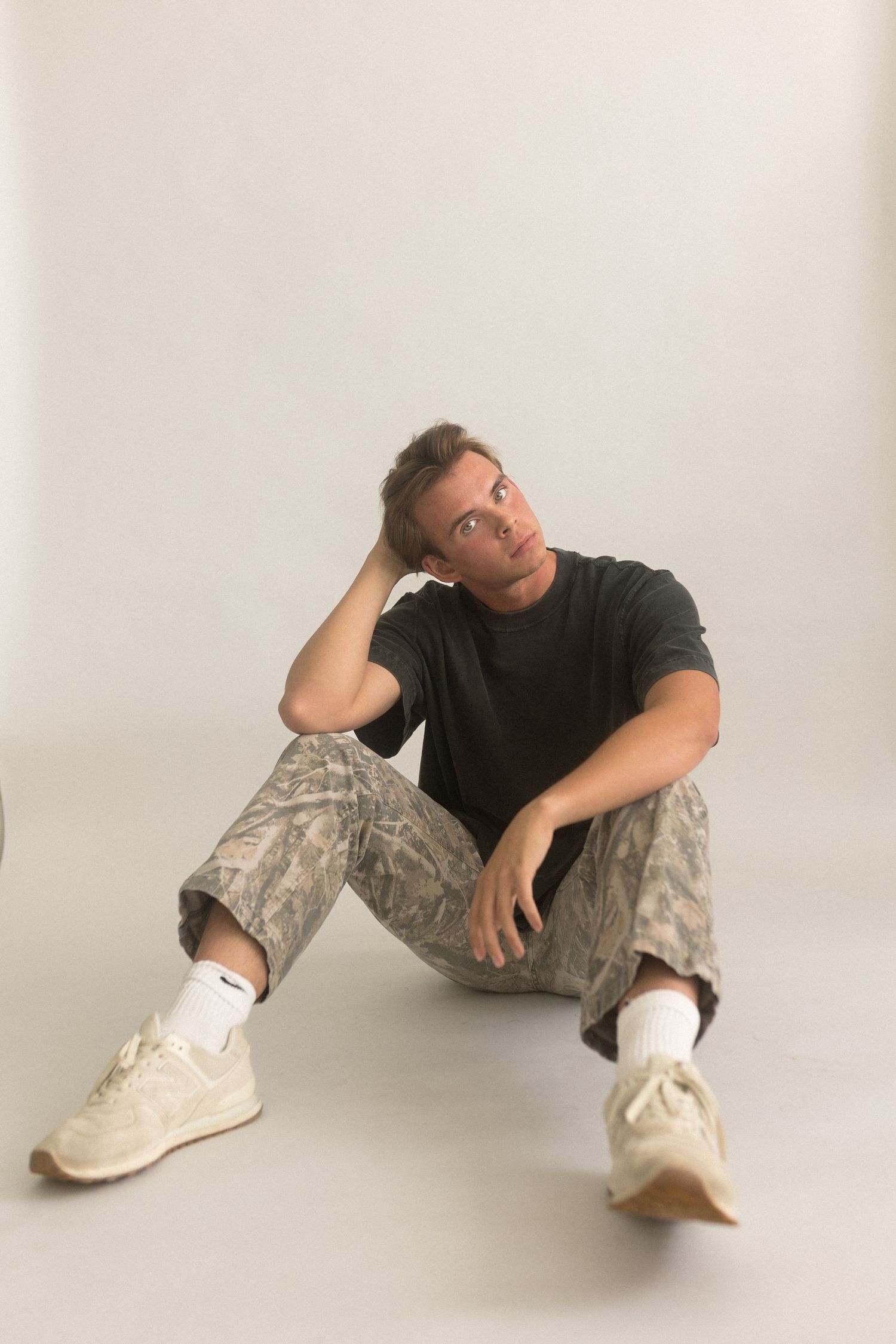 Model poses casually on floor wearing black t-shirt and camo pants against white backdrop.