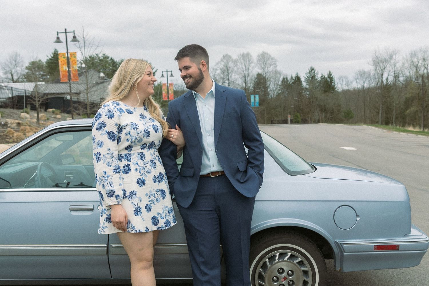 Joyful post-proposal moment, modern couple laughing in love beside a vintage car, candid engagement session in spring tones.