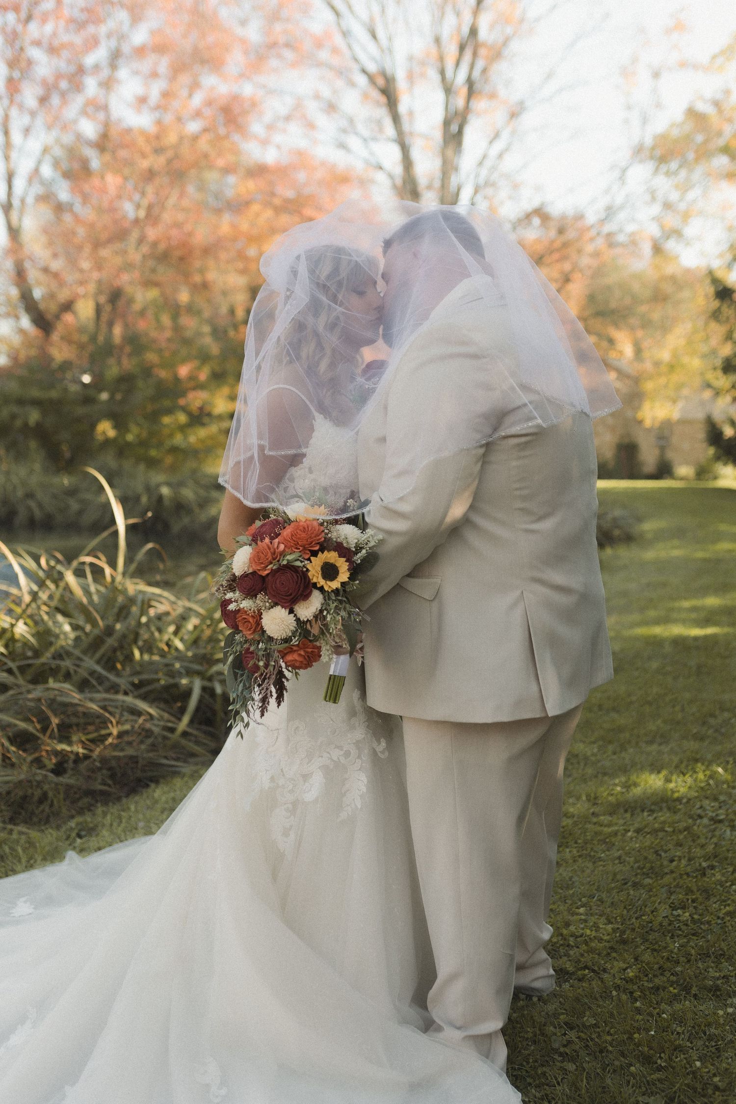 Newlyweds in light colored attire embrace while holding autumn bouquet outdoors.