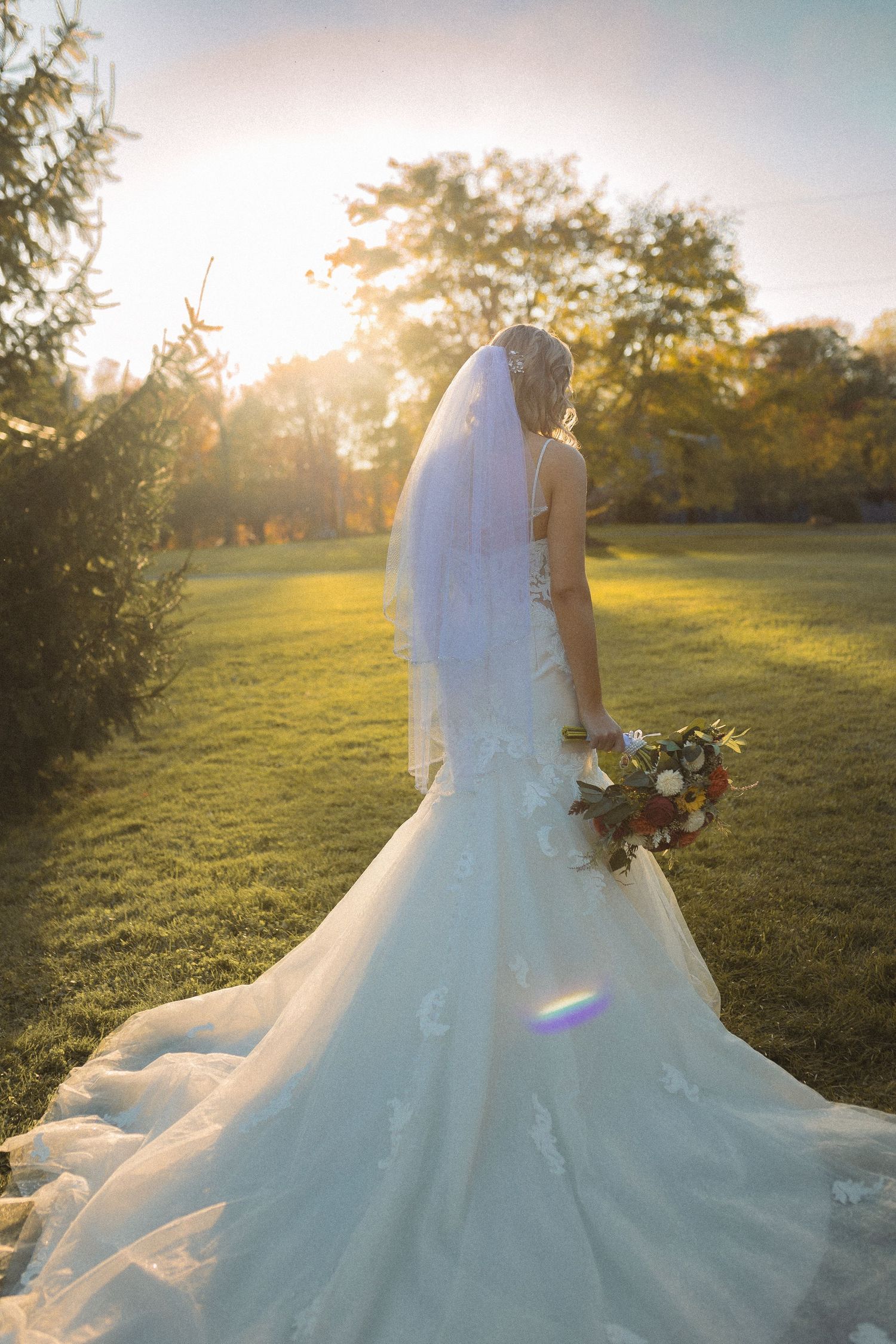 Bride in flowing white dress holds bouquet during golden hour in garden setting.